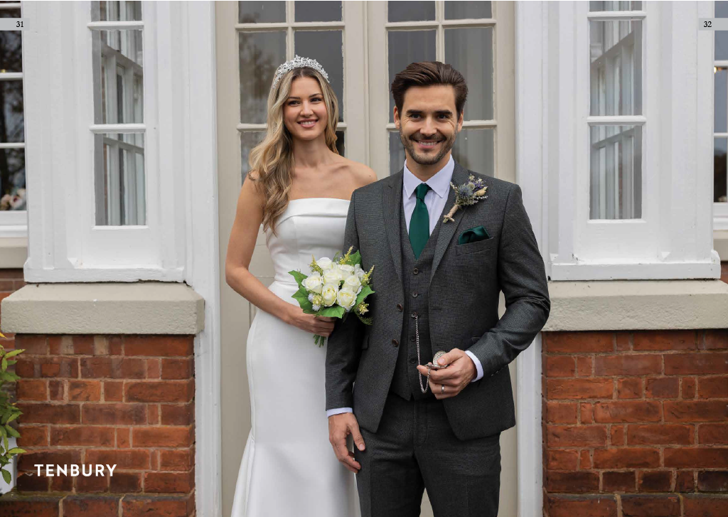 A bride and groom stand outside a building with white-framed windows, smiling for a wedding photo. The bride wears a strapless white wedding dress and a tiara, holding a bouquet of white roses. The groom wears a dark gray suit and green tie, holding 