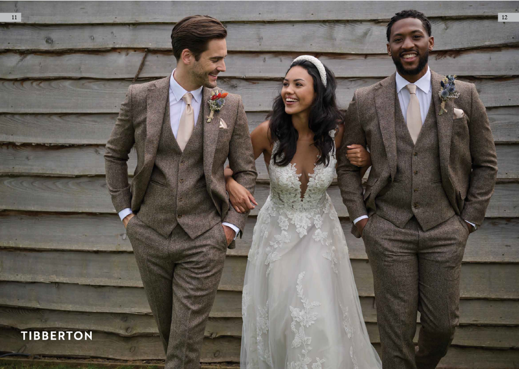 Three people dressed in wedding attire standing together in front of a wooden wall, smiling and enjoying each other's company. One woman in a white wedding gown is flanked by two men in brown suits.