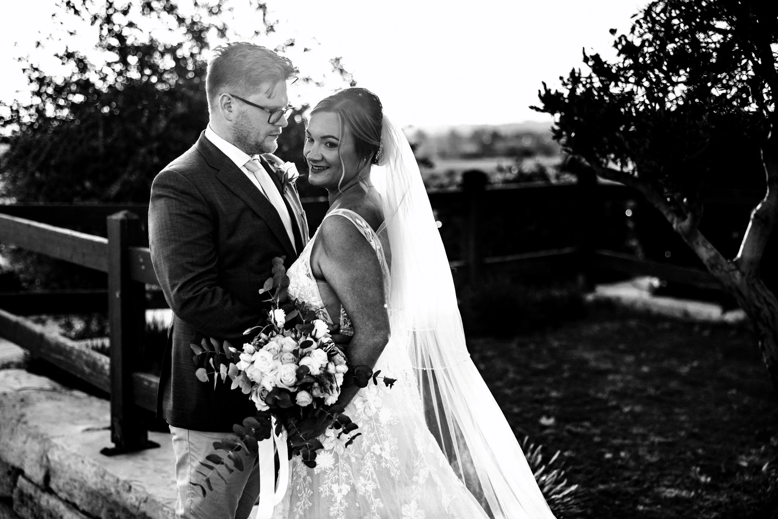 Groom gazes at bride while she looks to the side, her veil cascading down. Beautiful lace detail is highlighted by the sunlight in this black and white photo.