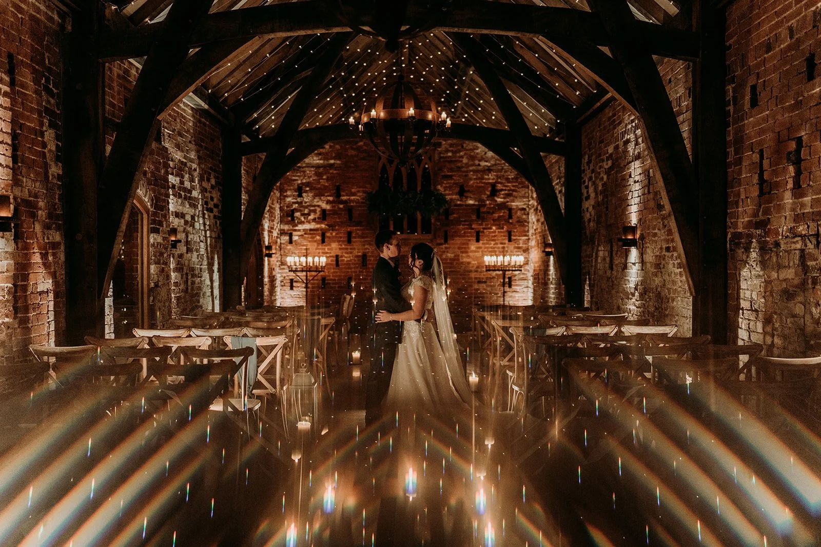 Festoon lights glow and sparkle in the ceremony room, which has the feel of a church. Candles frame the happy couple in the centre.