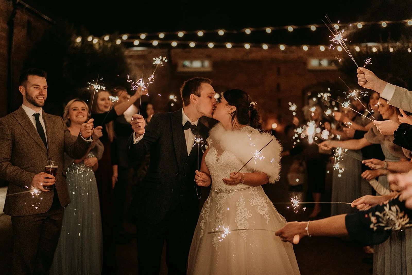The couple kiss surrounded by guests and sparklers. Festoon lights glow in the background.