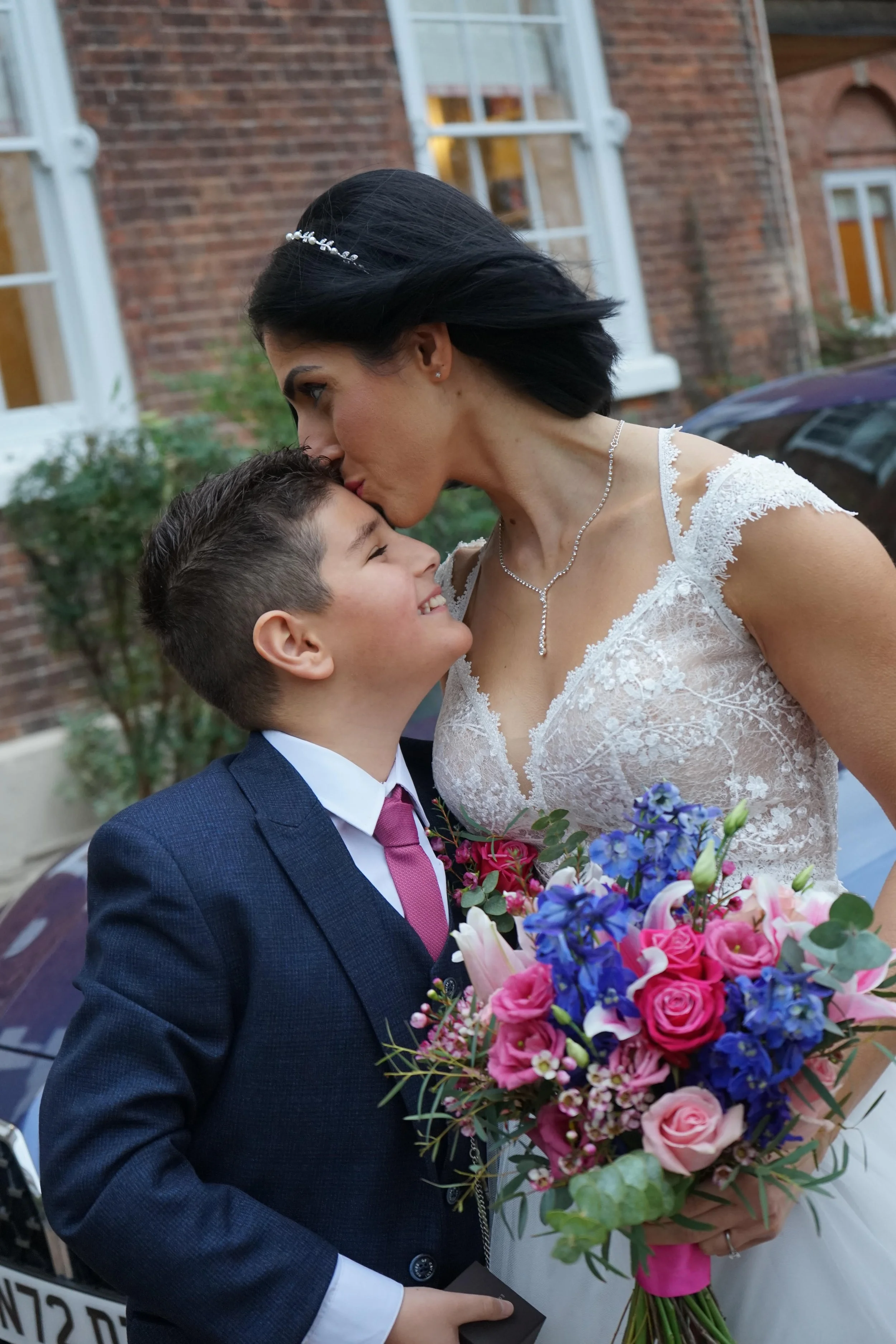 Motherly love on a special day @CarrieAustinPhotography. The bride kisses her son on the forehead. They are outside and the son is wearing a navy suit and pink tie.