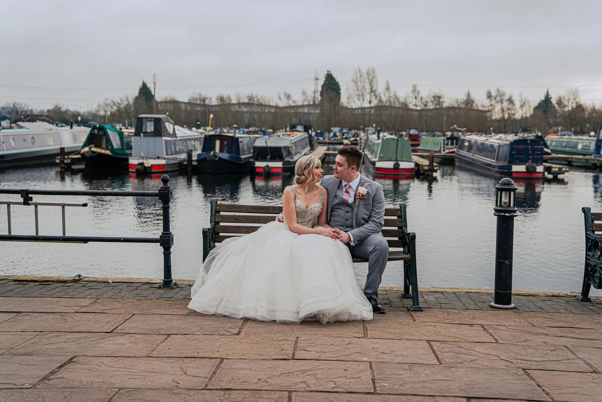 With the backdrop of boats reminiscent of their proposal, bride and groom sit on a bench looking lovingly at each other. @KeraRobson