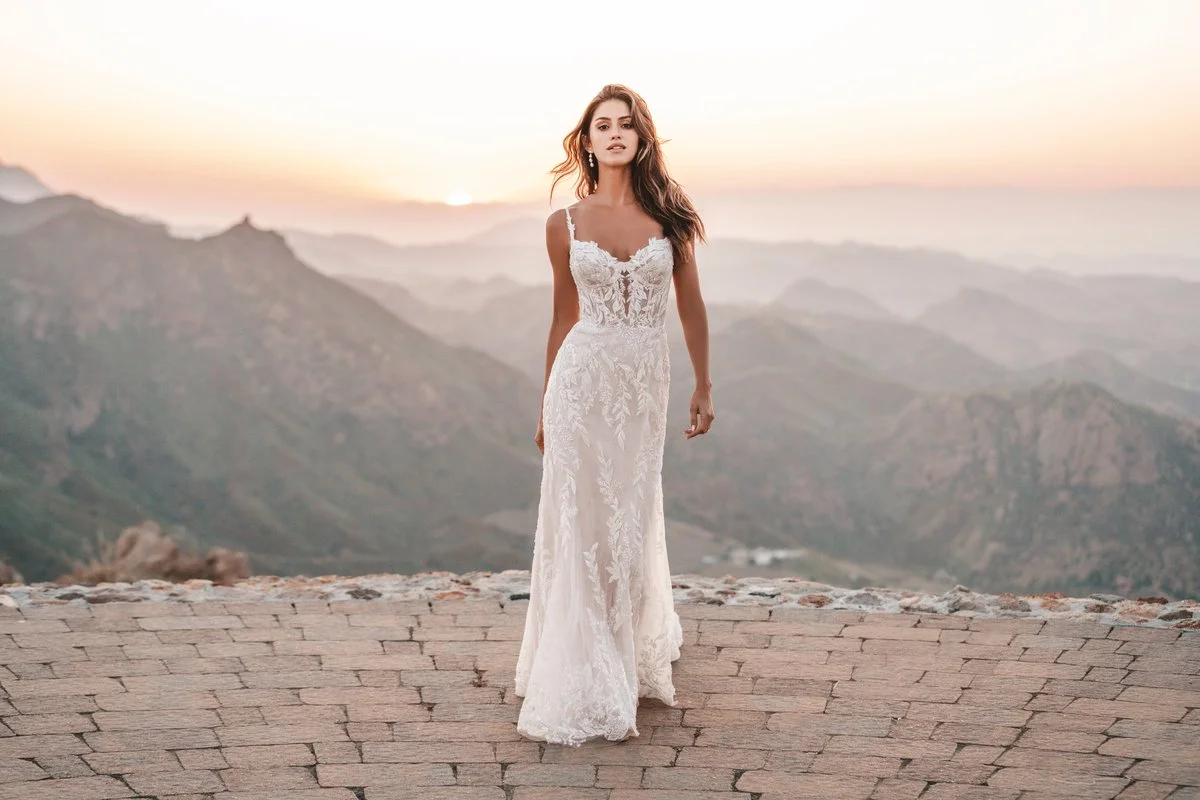 A woman in a white lace wedding dress standing on a stone platform with a mountainous landscape in the background at sunset.