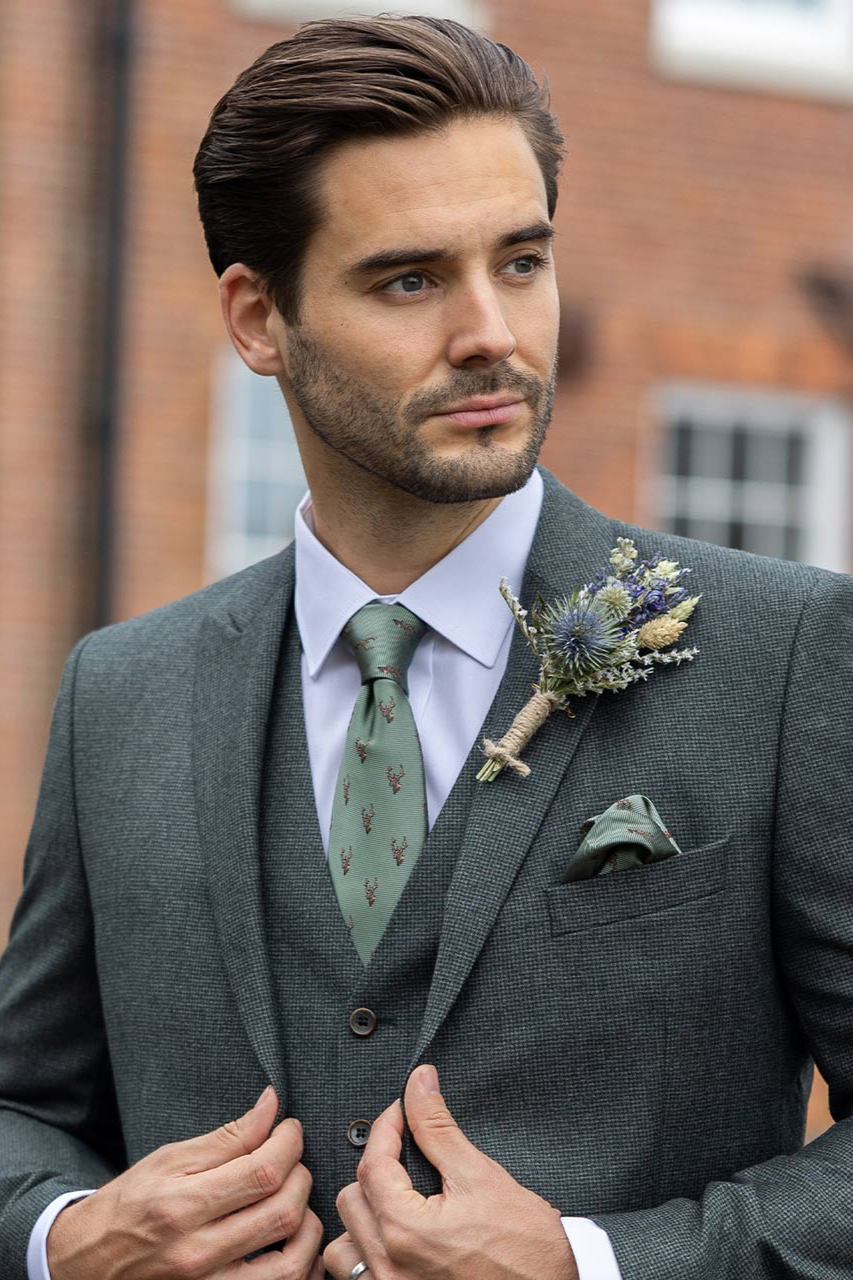 A man in a gray suit with a green tie and boutonniere stands outdoors, adjusting his suit jacket with both hands. Background features a blurred red-brick building.