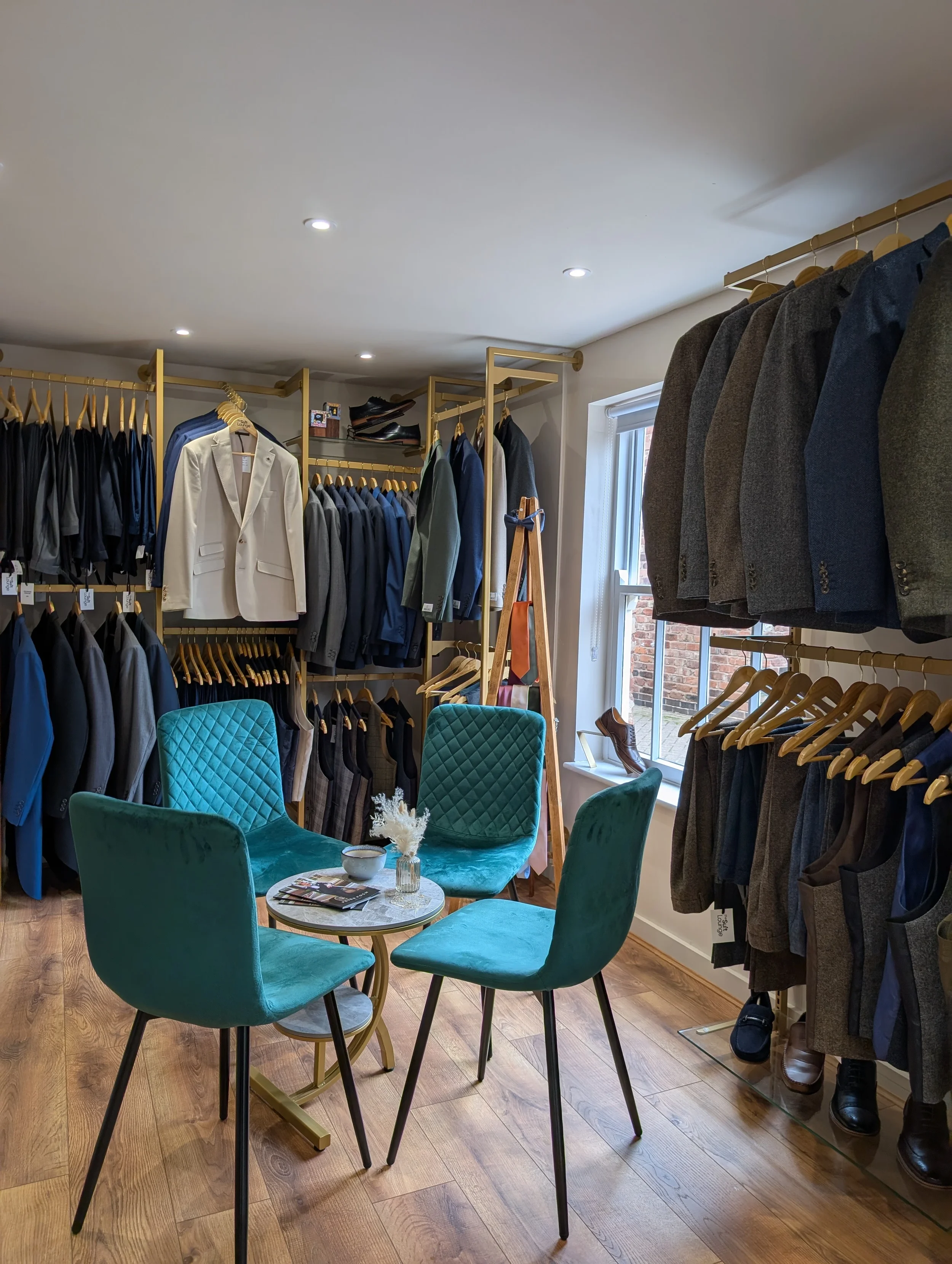 A boutique clothing store with clothing racks filled with suits and blazers, a small seating area with teal chairs and a marble table, and a window letting in natural light.