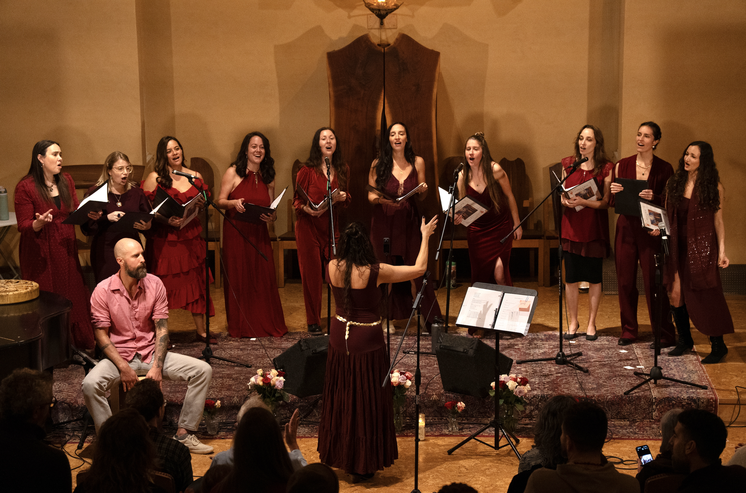 A women choir in red dresses singing on stage, with a conductor in front, in an indoor concert hall. The stage is decorated with pink and white flowers, and audience members are visible in the foreground.