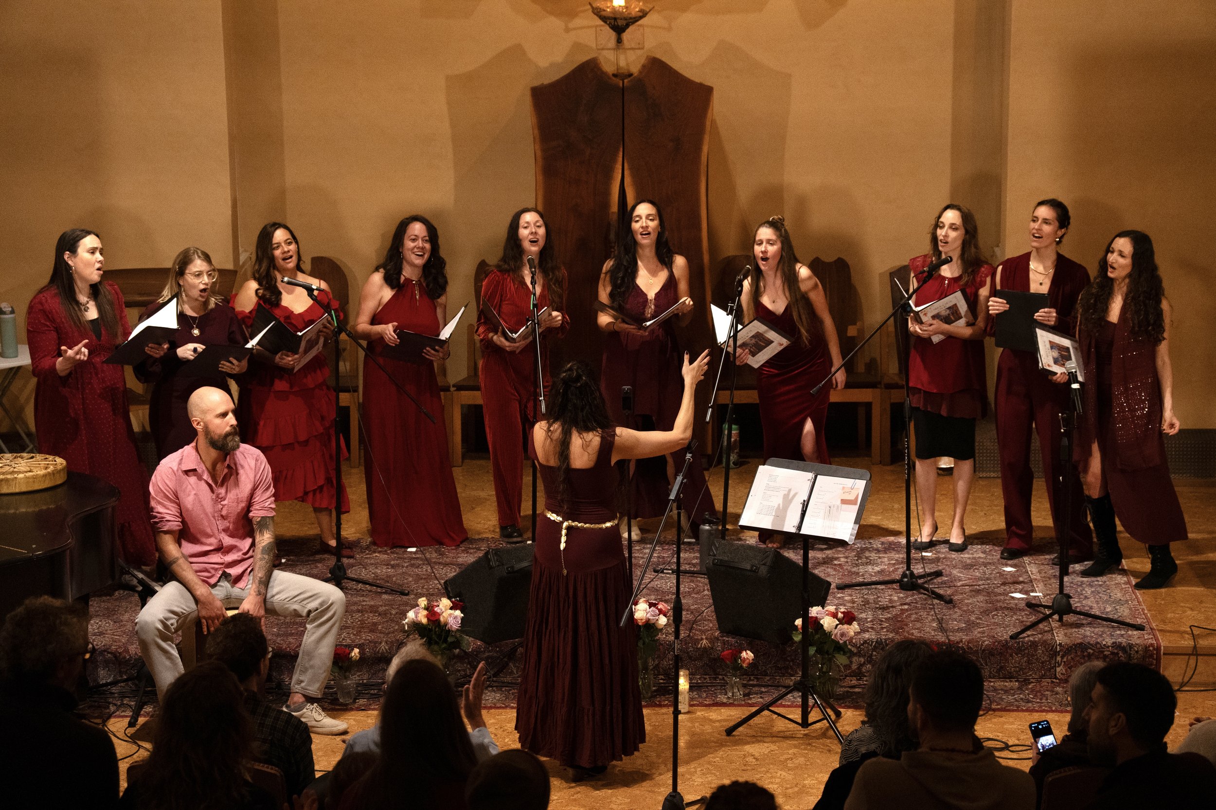 A choir of women dressed in red performing on stage with a conductor in front, and a man with a beard seated nearby, during a concert or performance.