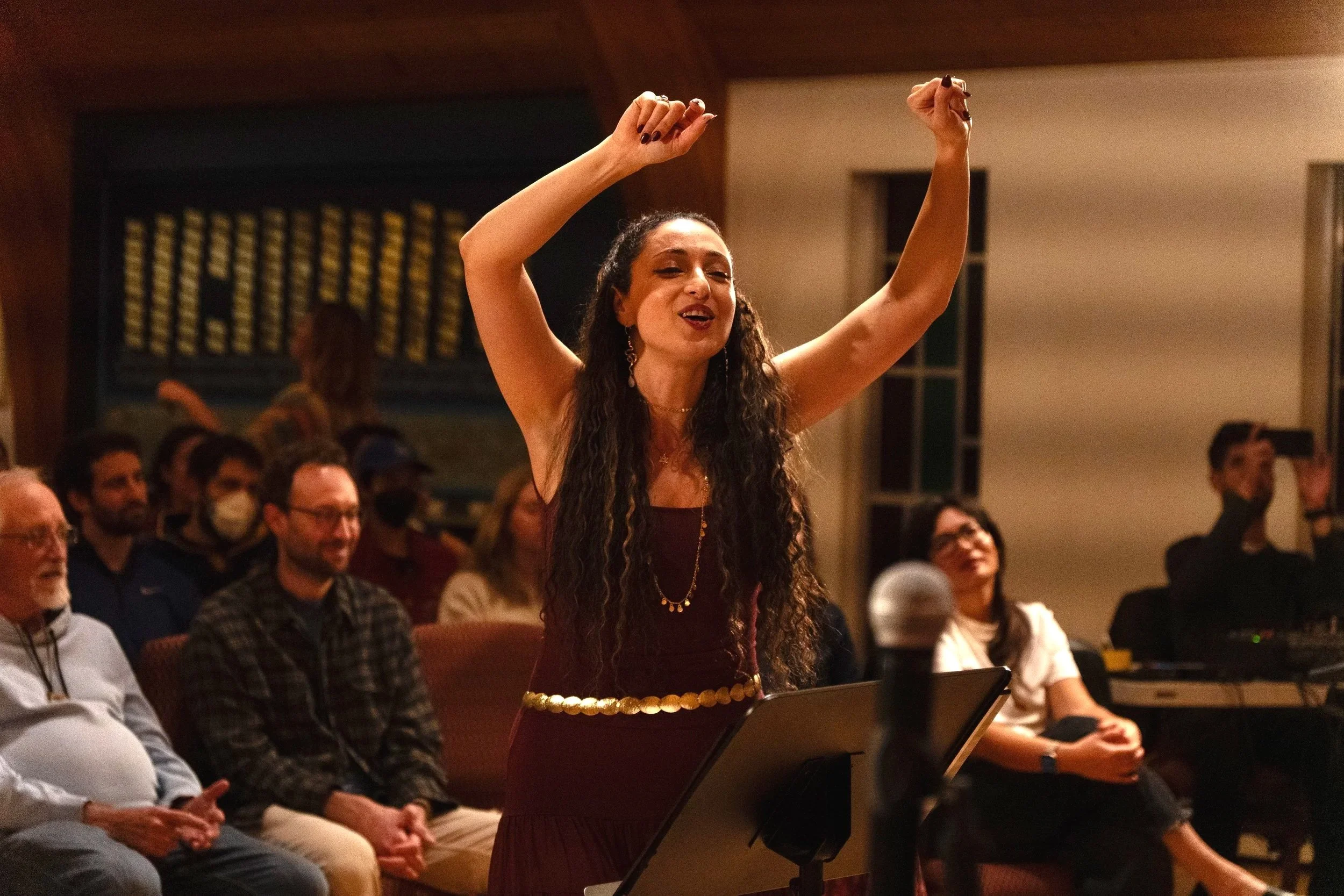 A woman with long curly hair dance lifts her arms up in a lively room filled with seated audience members.