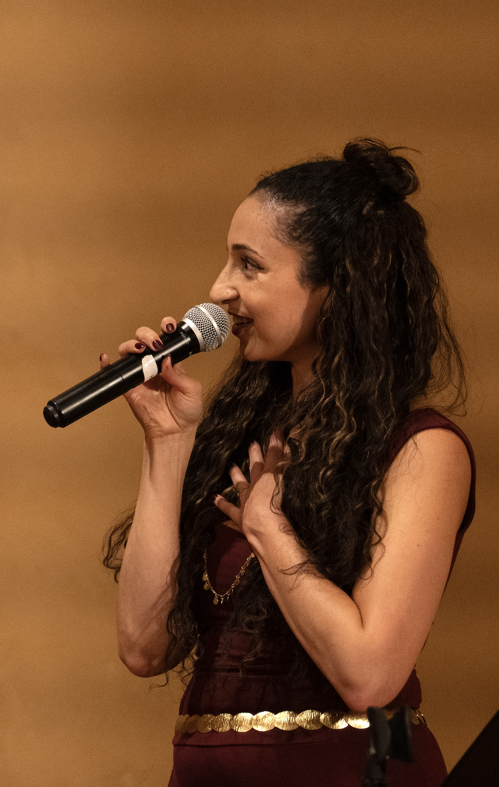 A woman with long curly hair, wearing a maroon dress with a gold belt, holding a microphone near her mouth, standing against a plain brown background.