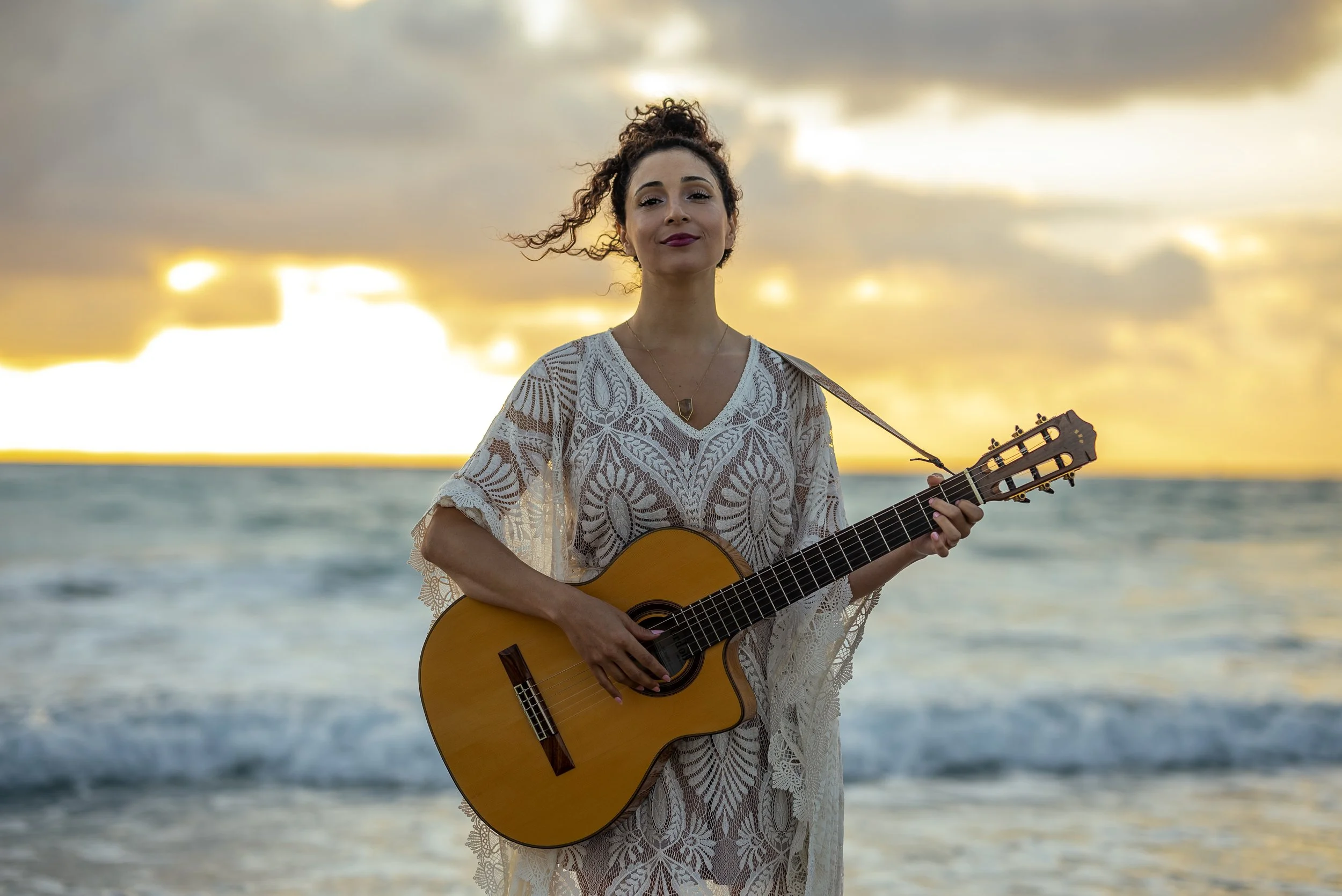 A woman with curly hair in a bun, wearing a white lace dress, stands on the beach holding an acoustic guitar against a sunset sky with clouds and ocean in the background.
