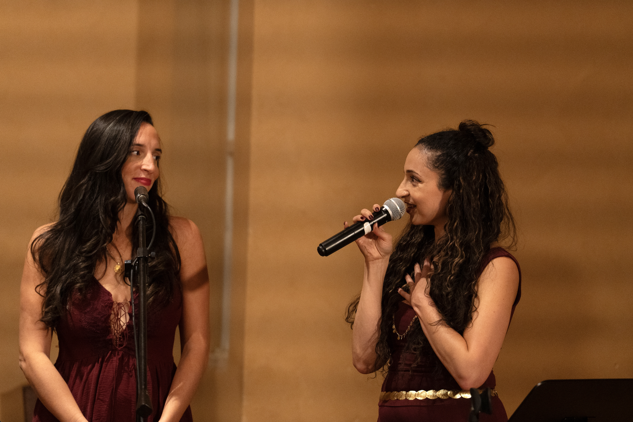 Two women dressed in elegant maroon dresses speaking or singing into microphones in a warm-colored room.