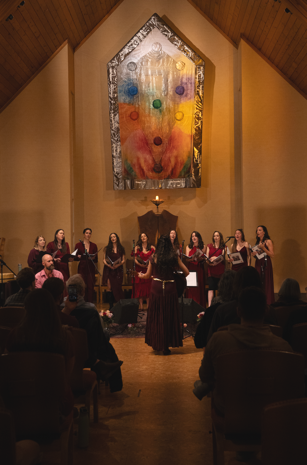 A choir of women wearing burgundy dresses performs on stage in a church, directed by a woman in a matching dress. The background features a colorful abstract stained glass window with geometric shapes and a candle holder with a lit candle. Audience members sit in chairs watching the performance.
