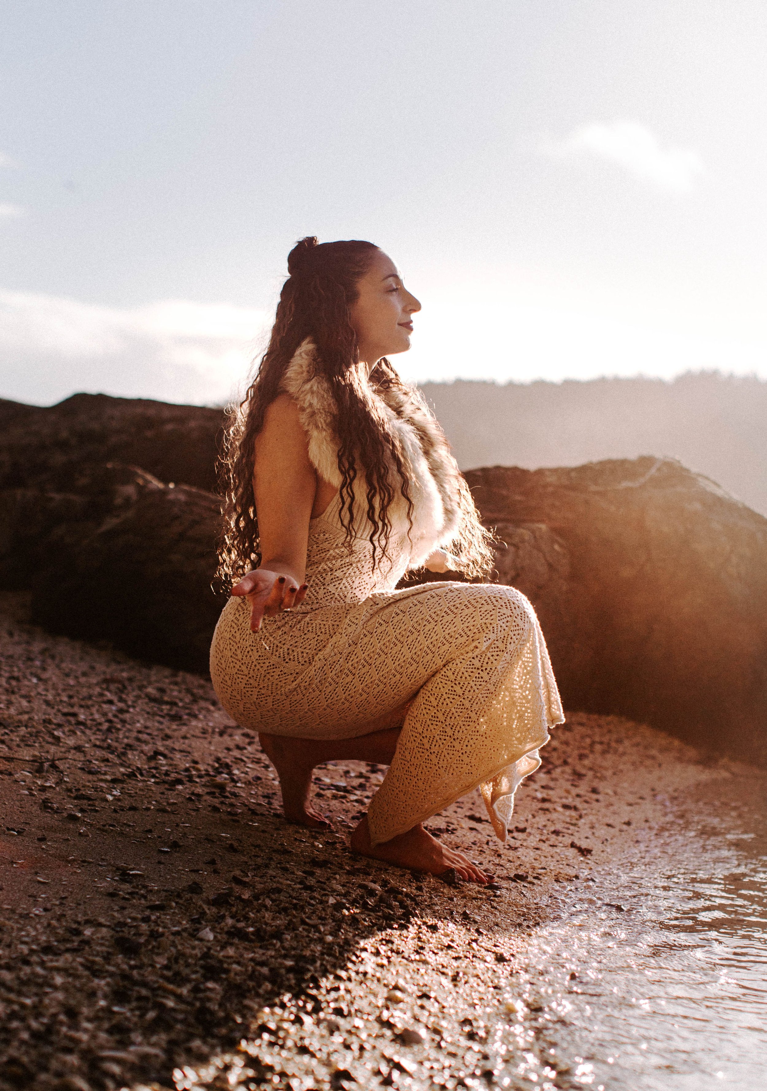 A woman with long curly hair and tan skin, sitting on a rocky beach at sunset, meditating with eyes closed and a serene expression, wearing a white crochet dress and a fur vest.