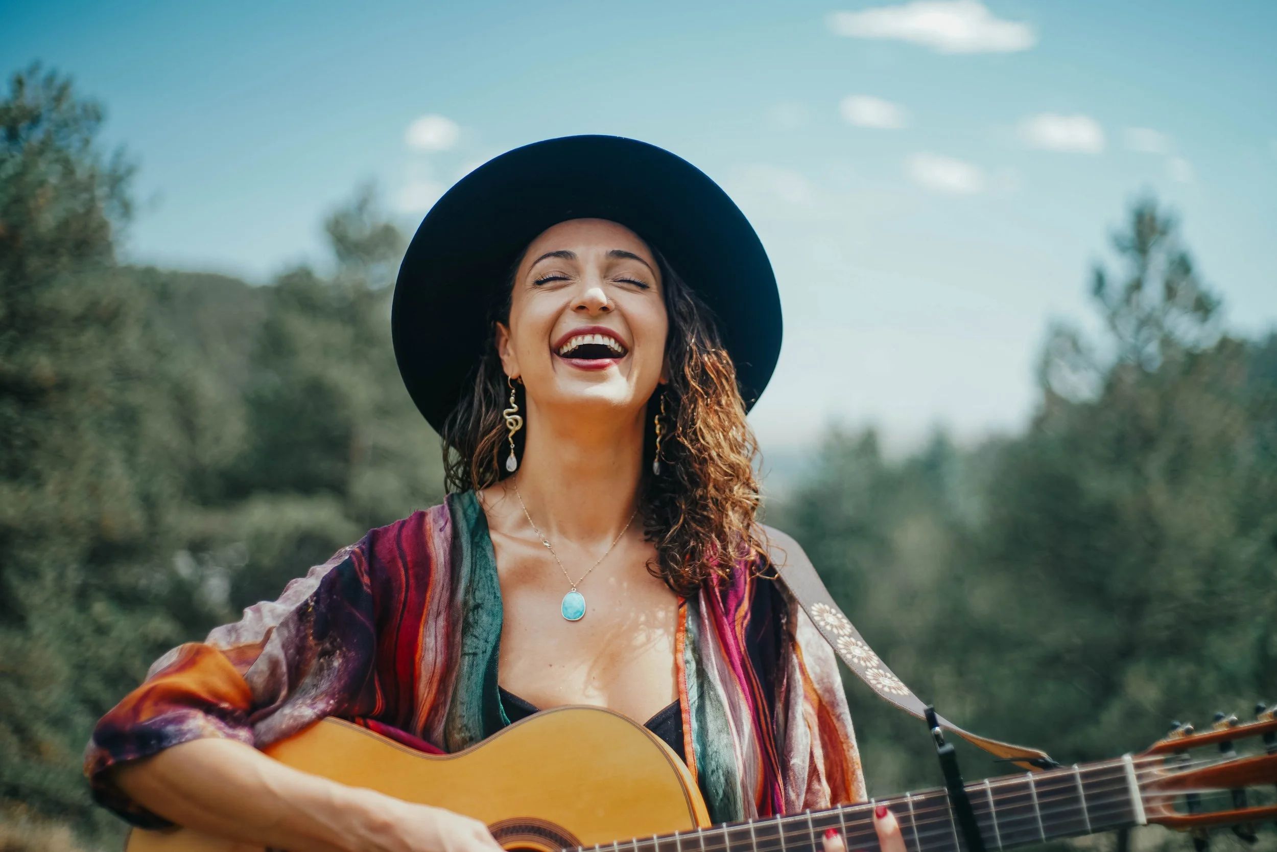 Woman with long curly hair wearing a black wide-brimmed hat, colorful dress, and jewelry, playing an acoustic guitar outdoors on a sunny day, smiling with eyes closed.
