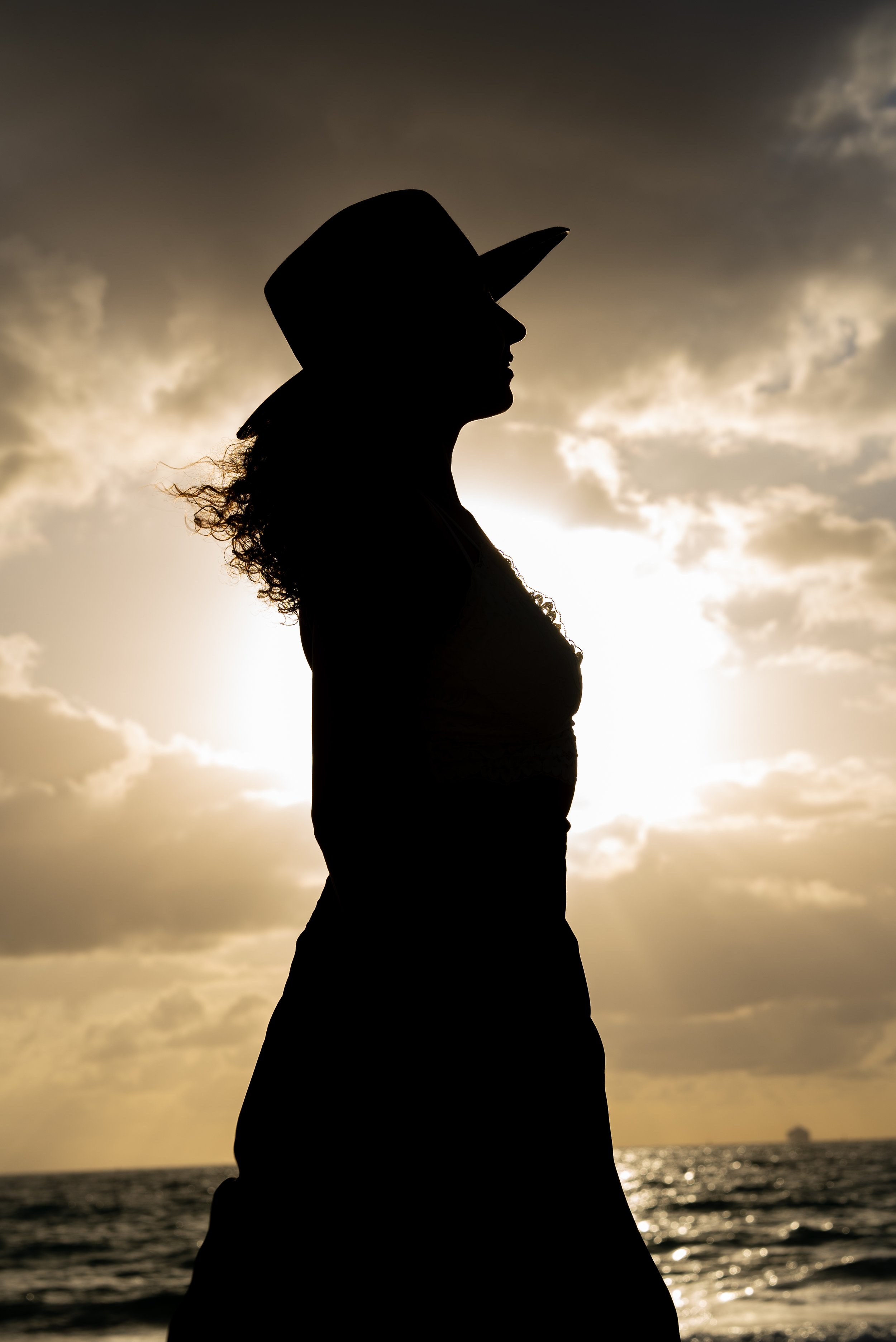 Silhouette of a woman wearing a cowboy hat standing by the water during sunset or sunrise, with clouds in the sky.