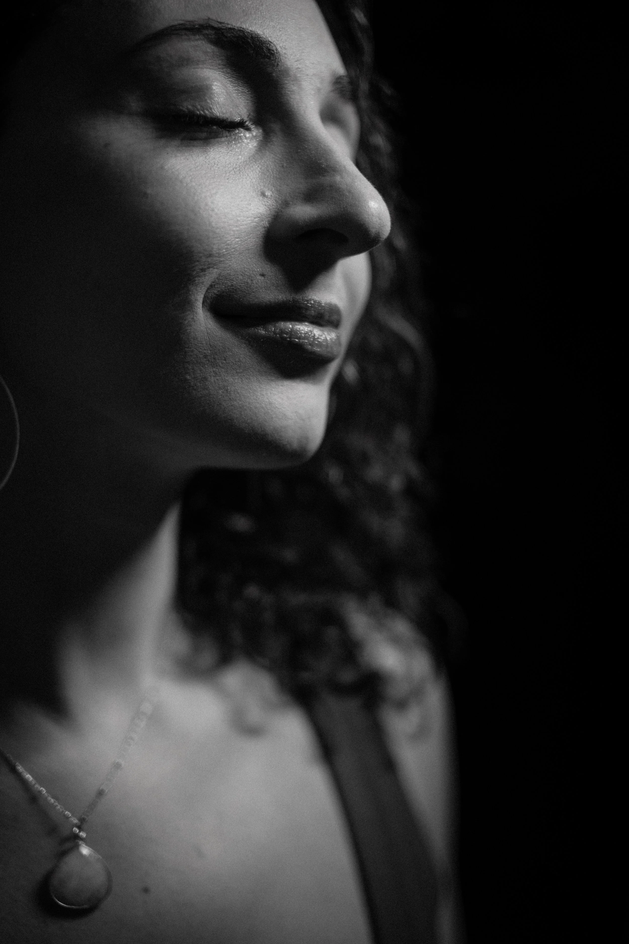 Close-up black and white photo of a woman's face with her eyes closed, smiling softly, wearing layered necklaces, with curly hair partially visible, against a dark background.
