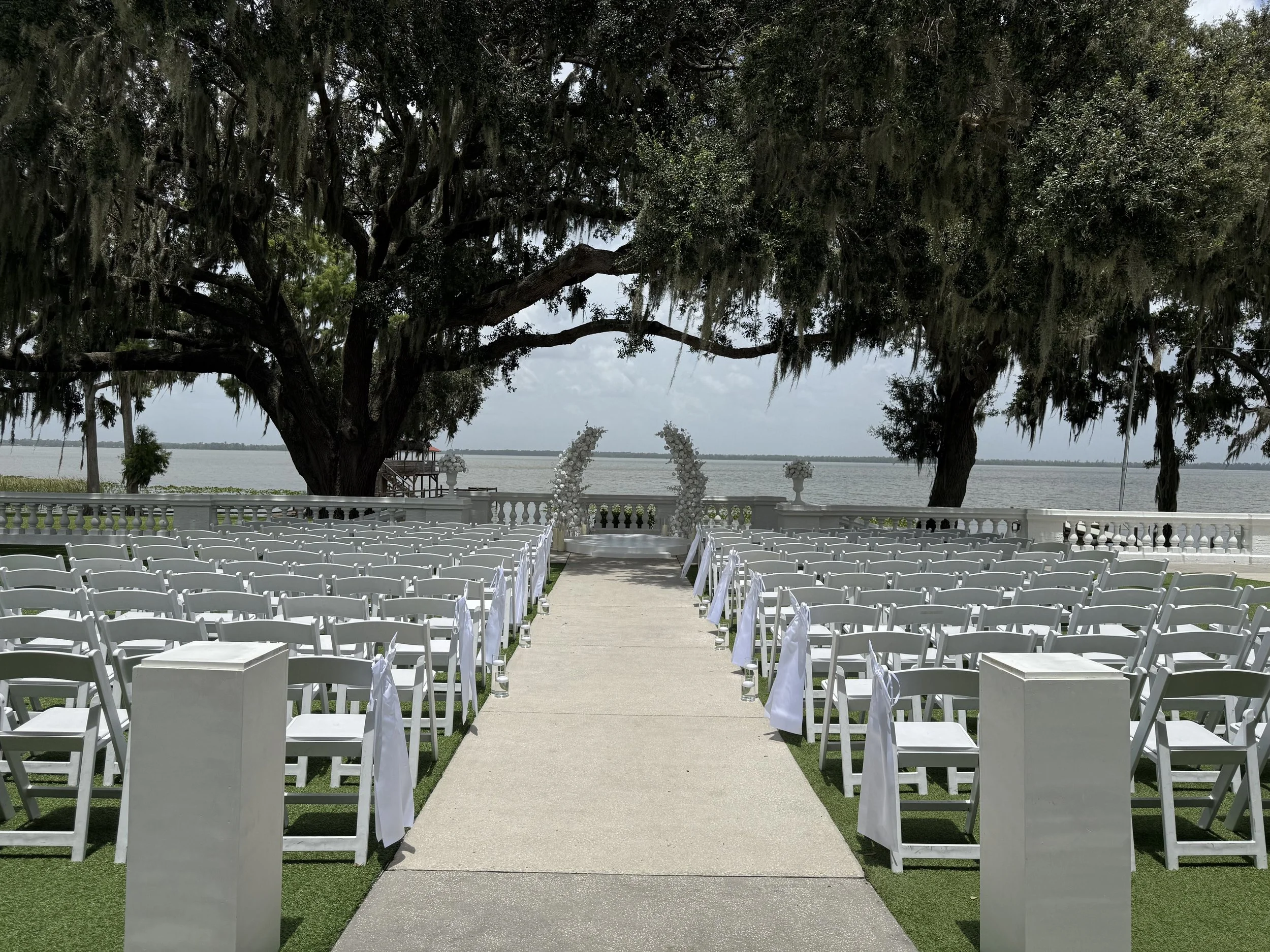 Outdoor wedding setup with white chairs lining a central aisle, decorated arch with white flowers, large trees in the background, and water beyond the trees.