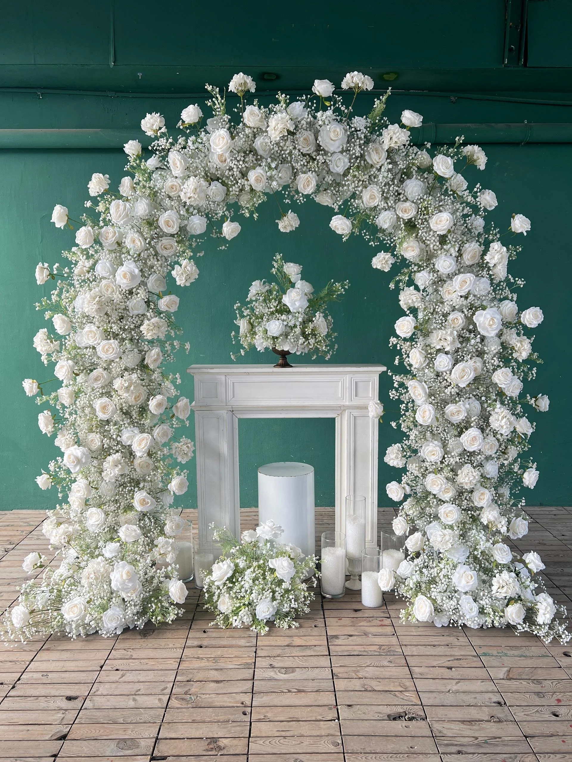 A white floral wedding arch decorated with roses and baby's breath positioned on a wooden deck against a green wall, with white candles in glass holders at the base.