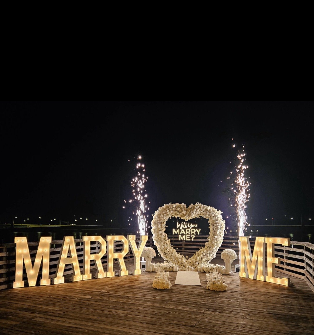 Outdoor night scene with large illuminated letters spelling 'MARRY' on the left and 'ME' on the right. In the center, decorative heart-shaped floral arrangement with the words 'Will you MARRY ME?' inside. Fireworks are exploding behind the setup, creating a celebratory atmosphere on a wooden deck near a body of water.