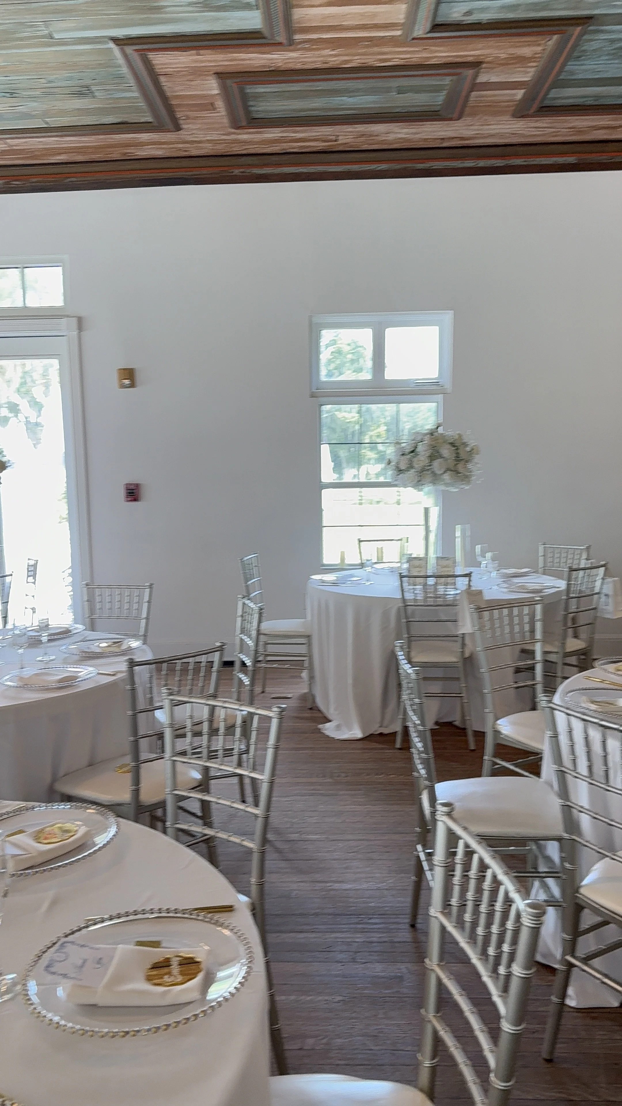 A banquet hall set up for a formal event with round tables covered in white tablecloths and arranged with place settings, silver Chiavari chairs, and floral centerpieces near the windows with natural light.