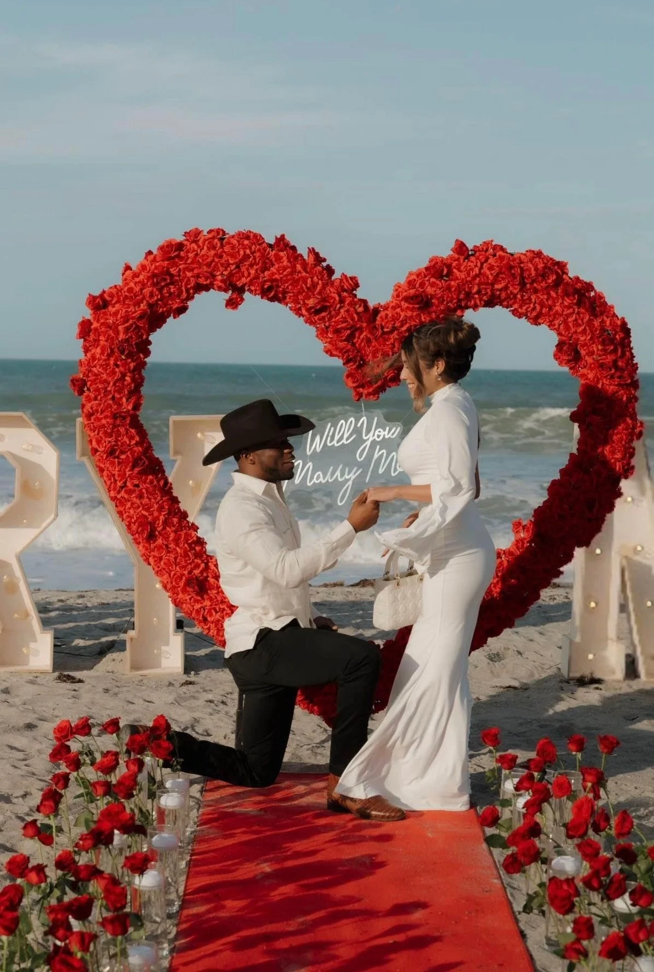 A couple gets engaged on a beach with a red heart-shaped floral arch and red roses lining a red carpet aisle.