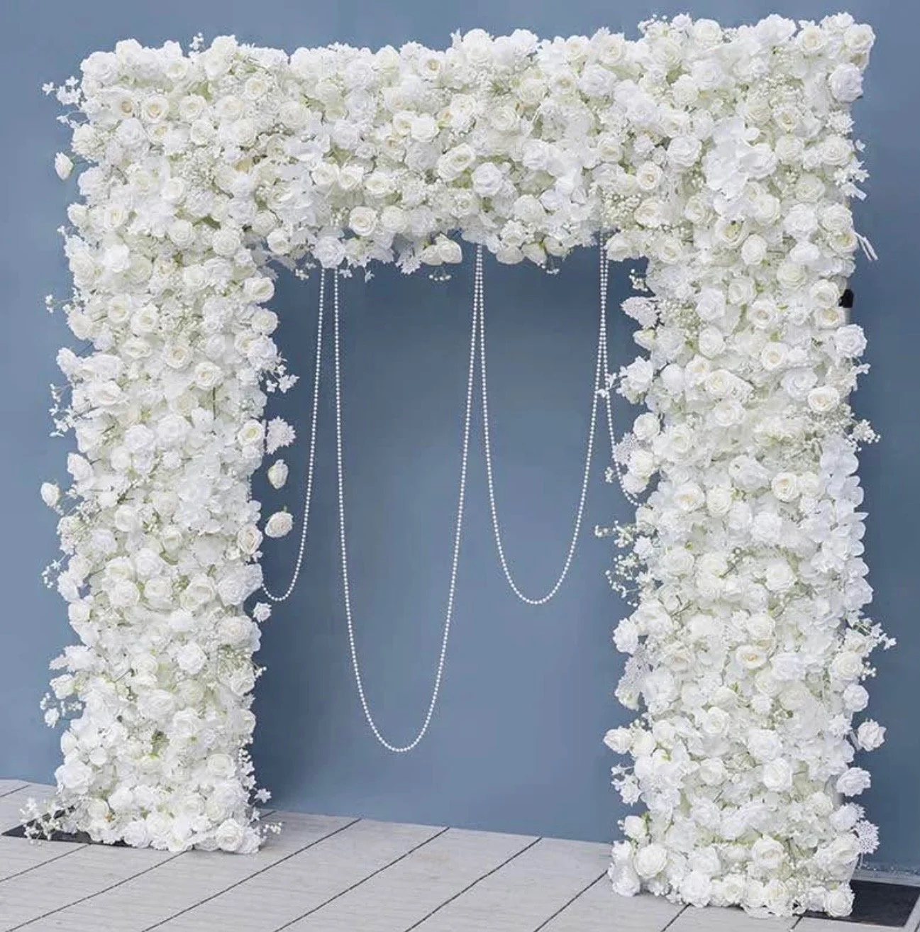 White floral wedding arch with roses and hanging pearl strings against a blue background.
