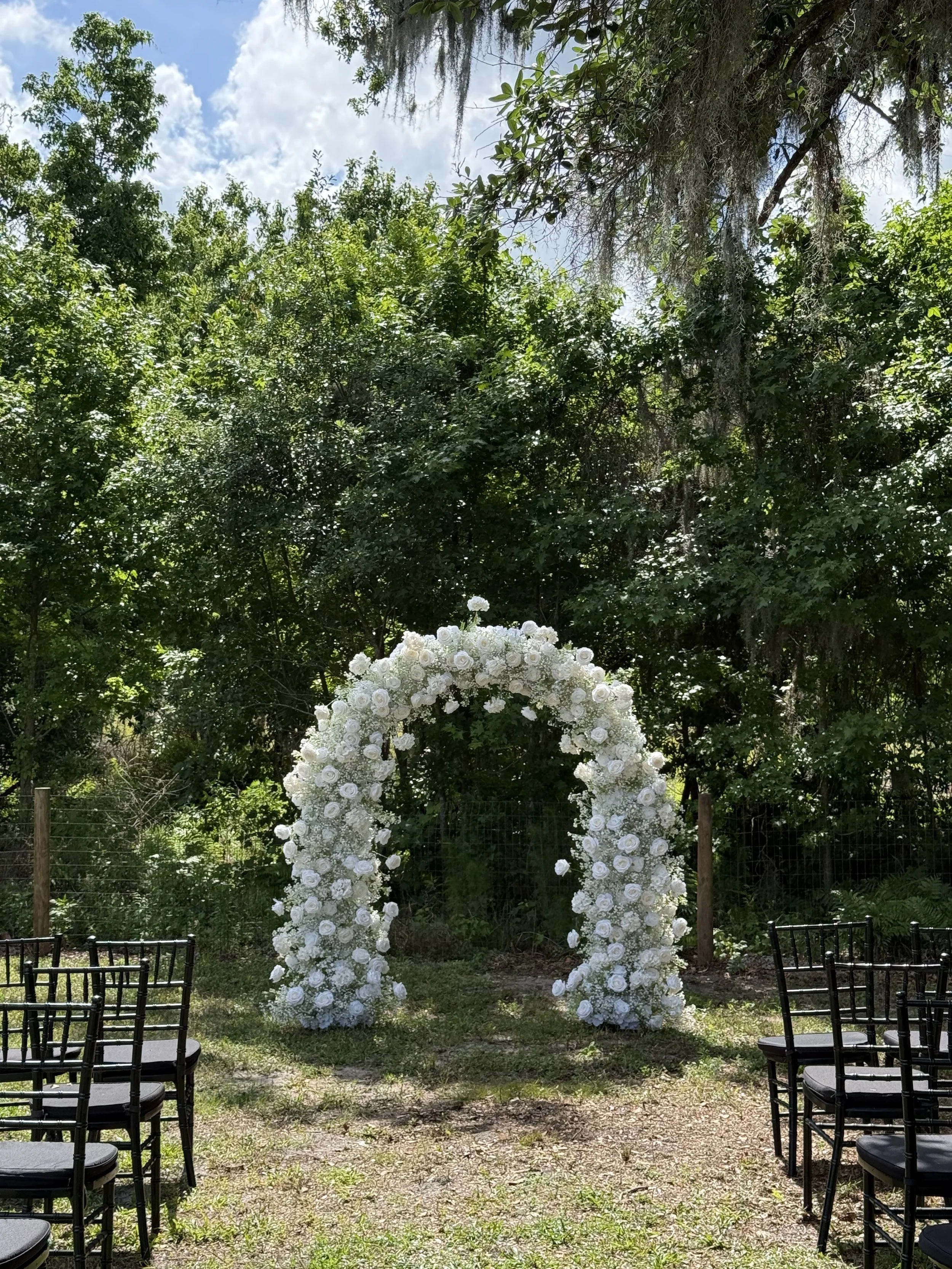 White floral wedding arch in an outdoor garden setting surrounded by trees and chairs for guests.