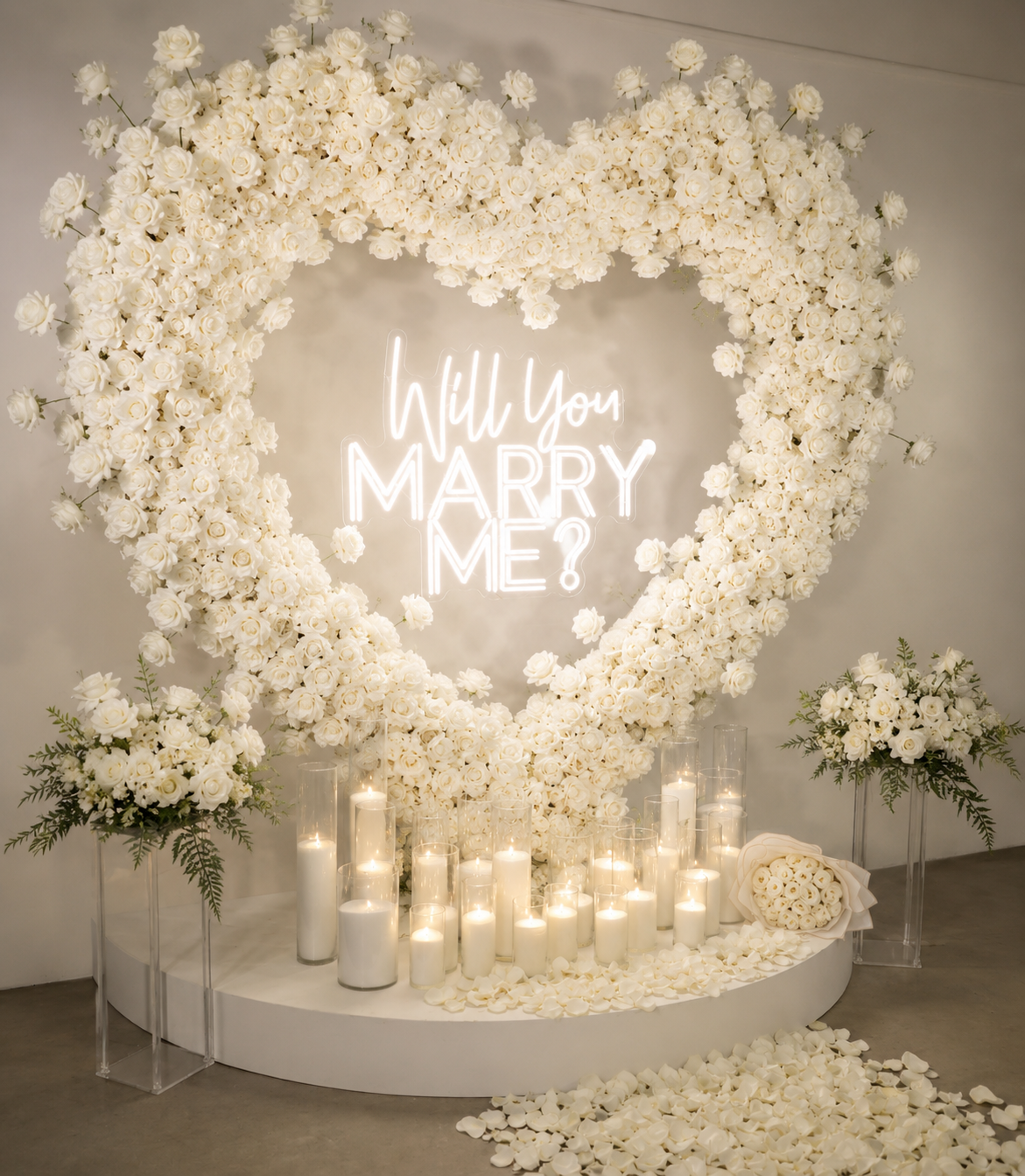 A romantic display with a heart-shaped floral wreath made of white roses, candles, and a neon sign that reads 'Will You Marry Me?' with additional smaller bouquet arrangements of white roses and greenery.
