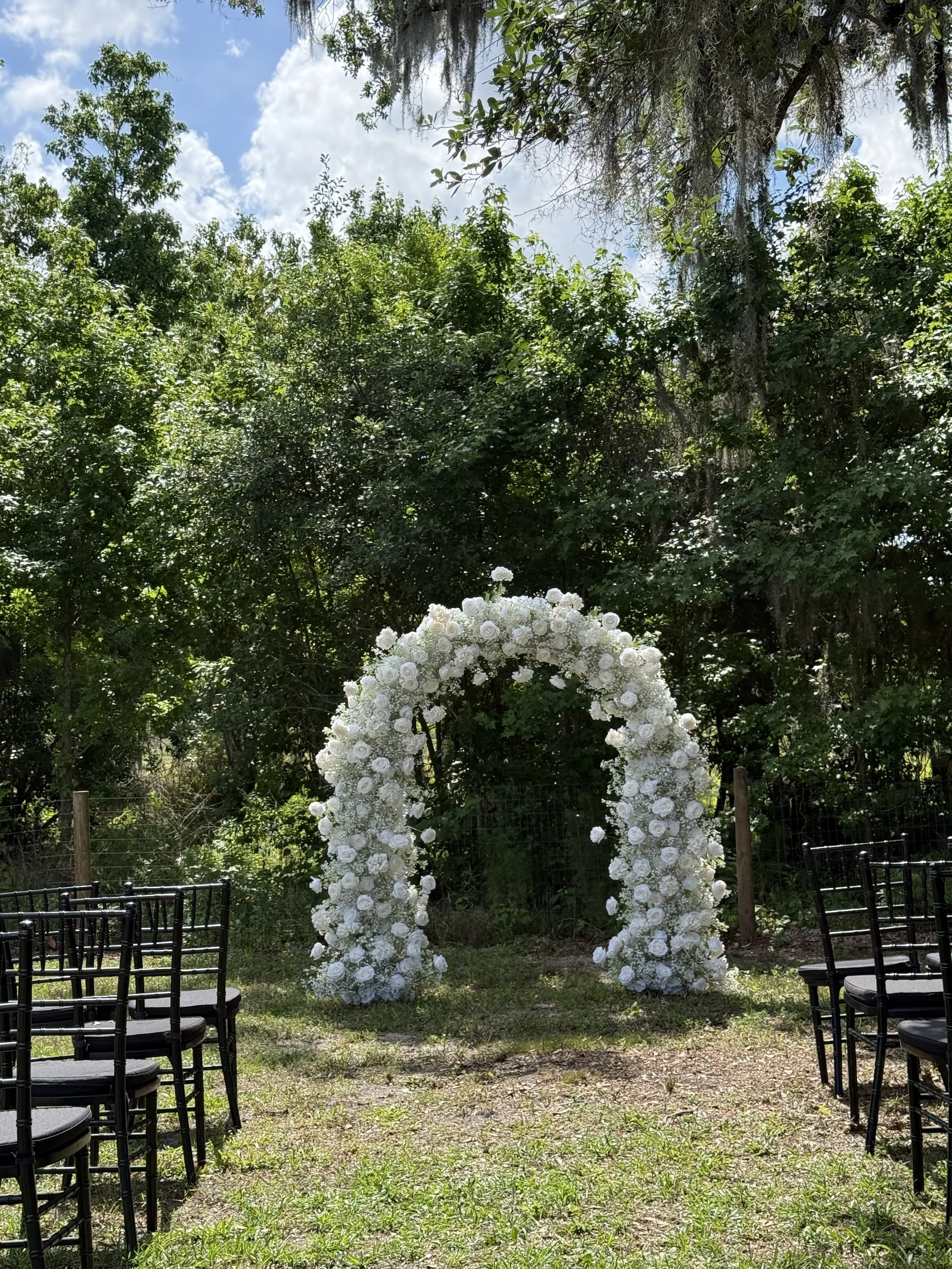 Outdoor wedding arch decorated with white roses in a lush green garden with chairs arranged on both sides.