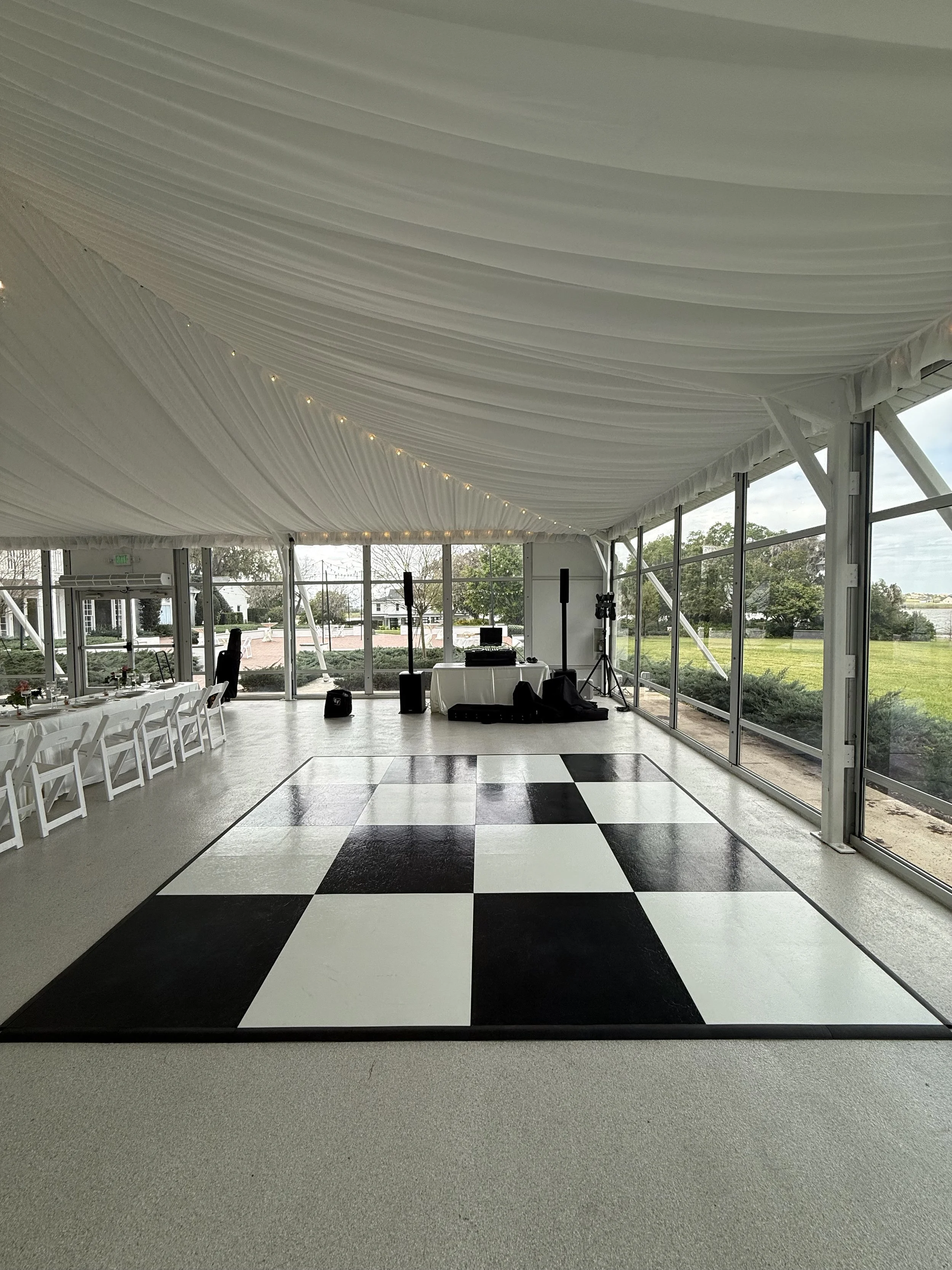Interior of a tented event space with black and white checkered dance floor, tables and chairs, and a DJ setup, with large windows showing outdoor greenery.