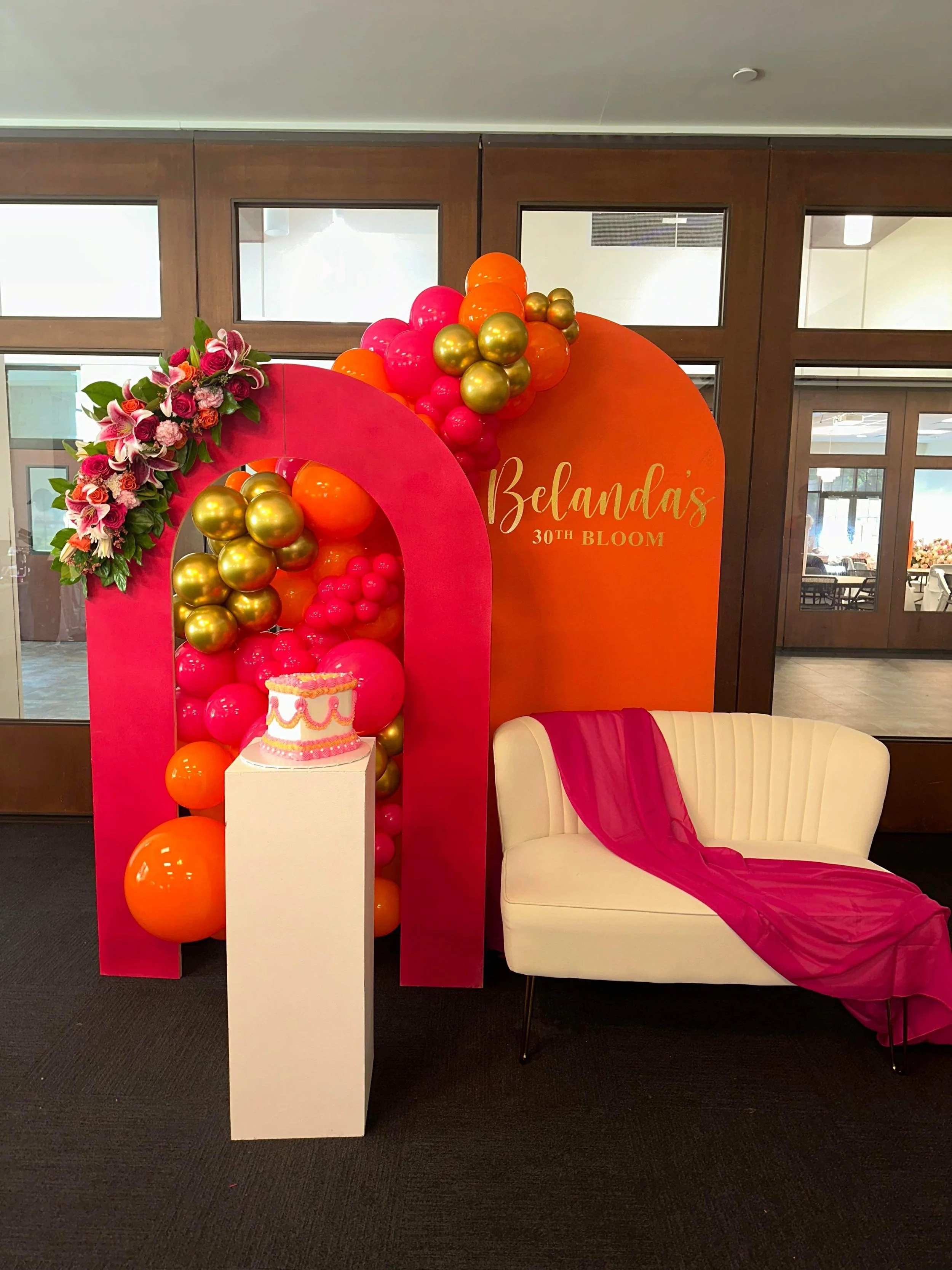 Decorative event display with balloons, flowers, a cake, and a sign reading 'Belanda's 30th Bloom,' featuring pink, gold, and orange colors, set inside a room with glass doors and windows.