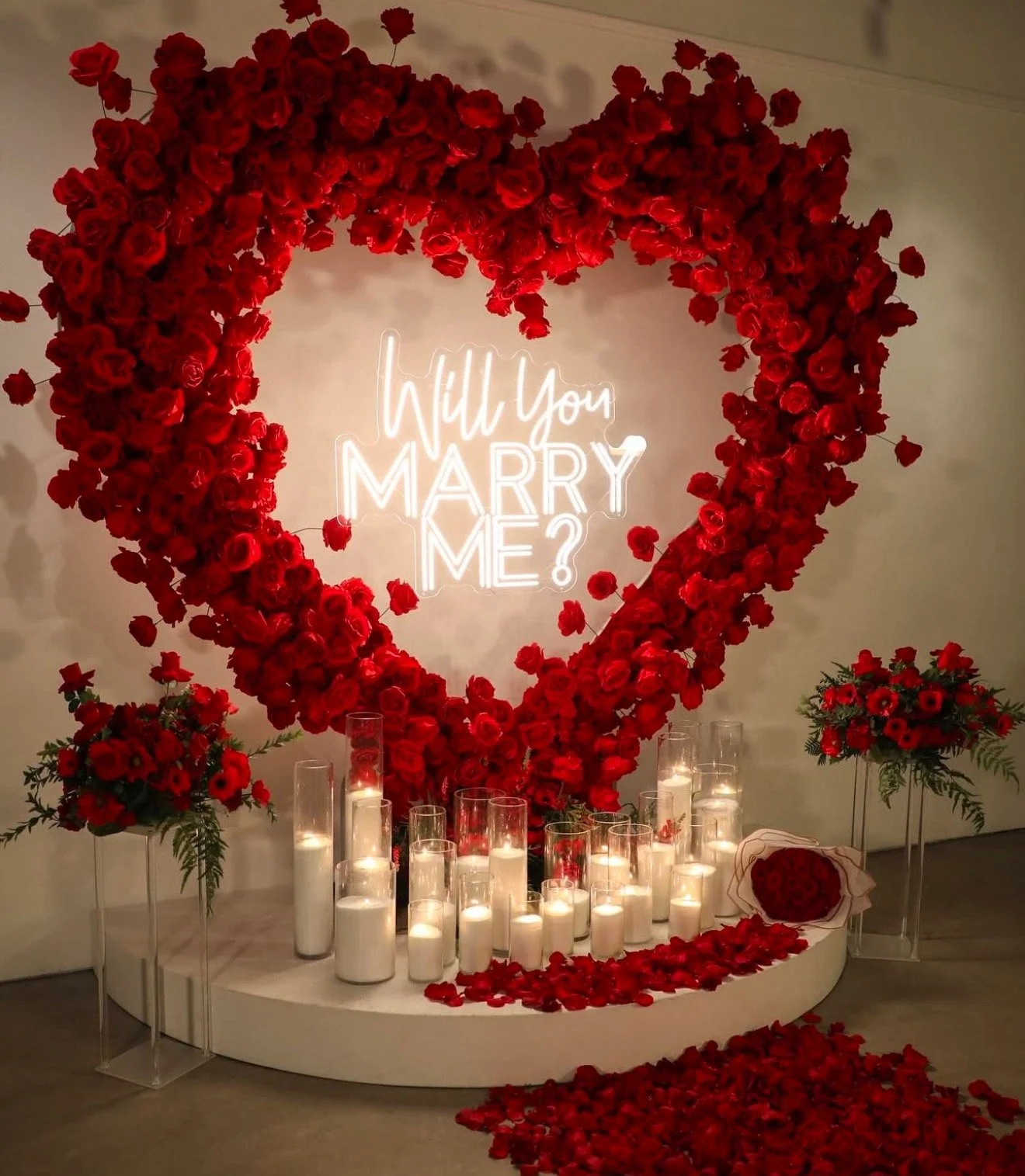 A romantic wedding proposal display featuring a large heart-shaped arrangement of red roses, illuminated sign with the words "Will You Marry Me?" in neon lights, surrounded by candles in tall glass holders and smaller bouquets of red flowers, with rose petals scattered on the floor.