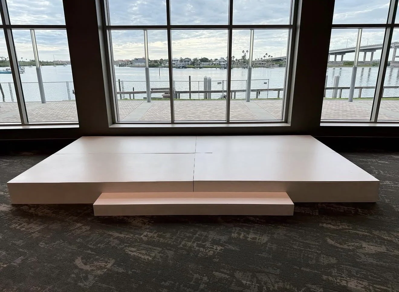 Empty white stage or platform inside a room with large windows overlooking a waterway and bridge.