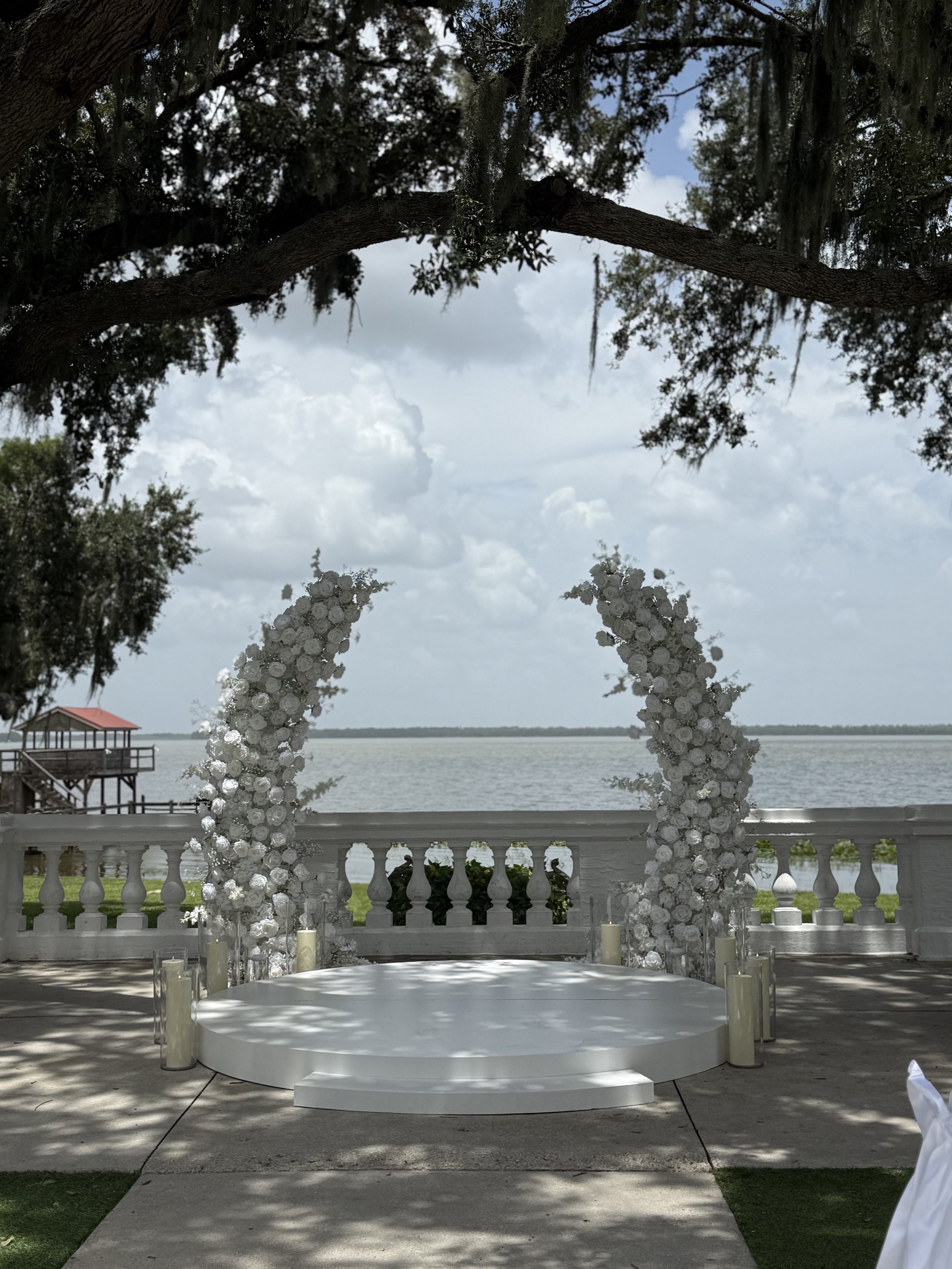 A circular white wedding altar decorated with white roses and candles, set outdoors near a body of water, with tree branches above and a cloudy sky in the background.
