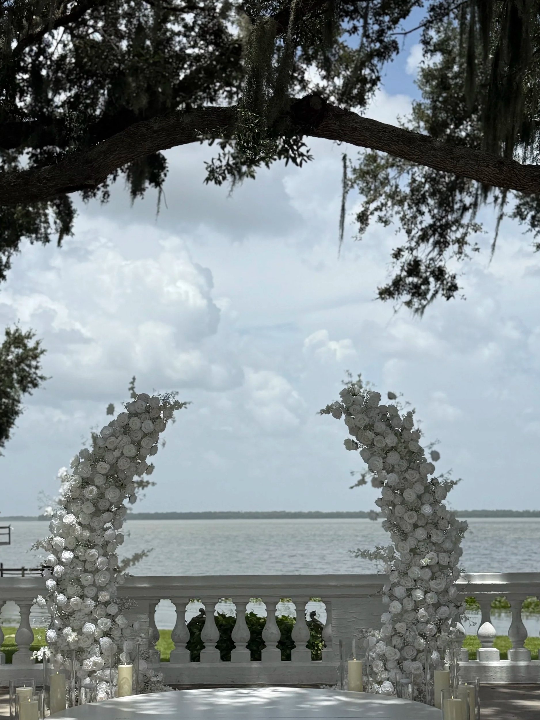 A scenic view of a lake with a decorative white railing in the foreground, framed by trees and clouds in the sky. The scene appears to be outdoors near a waterfront, with a cloudy sky overhead.