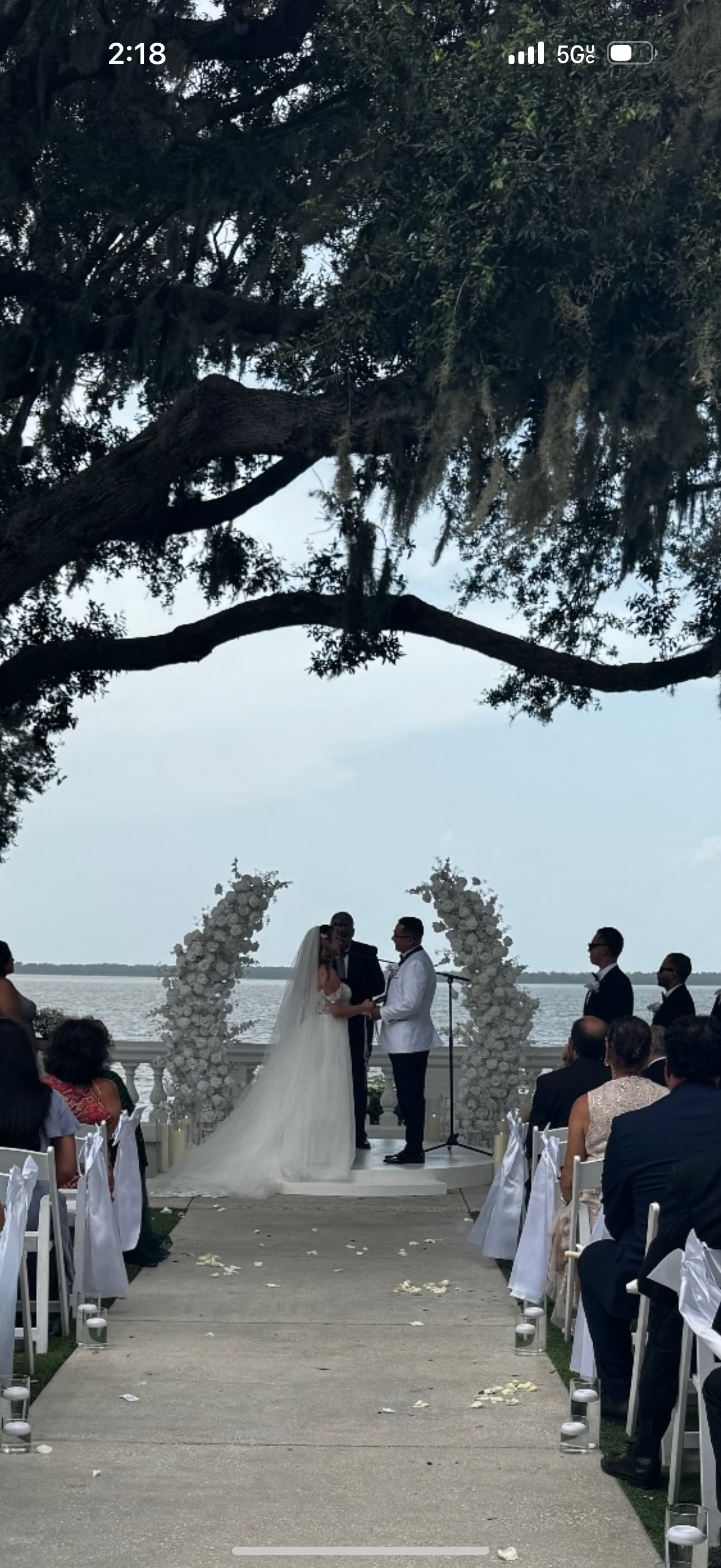 A wedding ceremony outdoors by a body of water, with the bride and groom holding hands in front of an officiant. Guests are seated on either side of the aisle, decorated with white ribbons and flowers, with large floral arrangements behind the couple and a tree overhanging above.