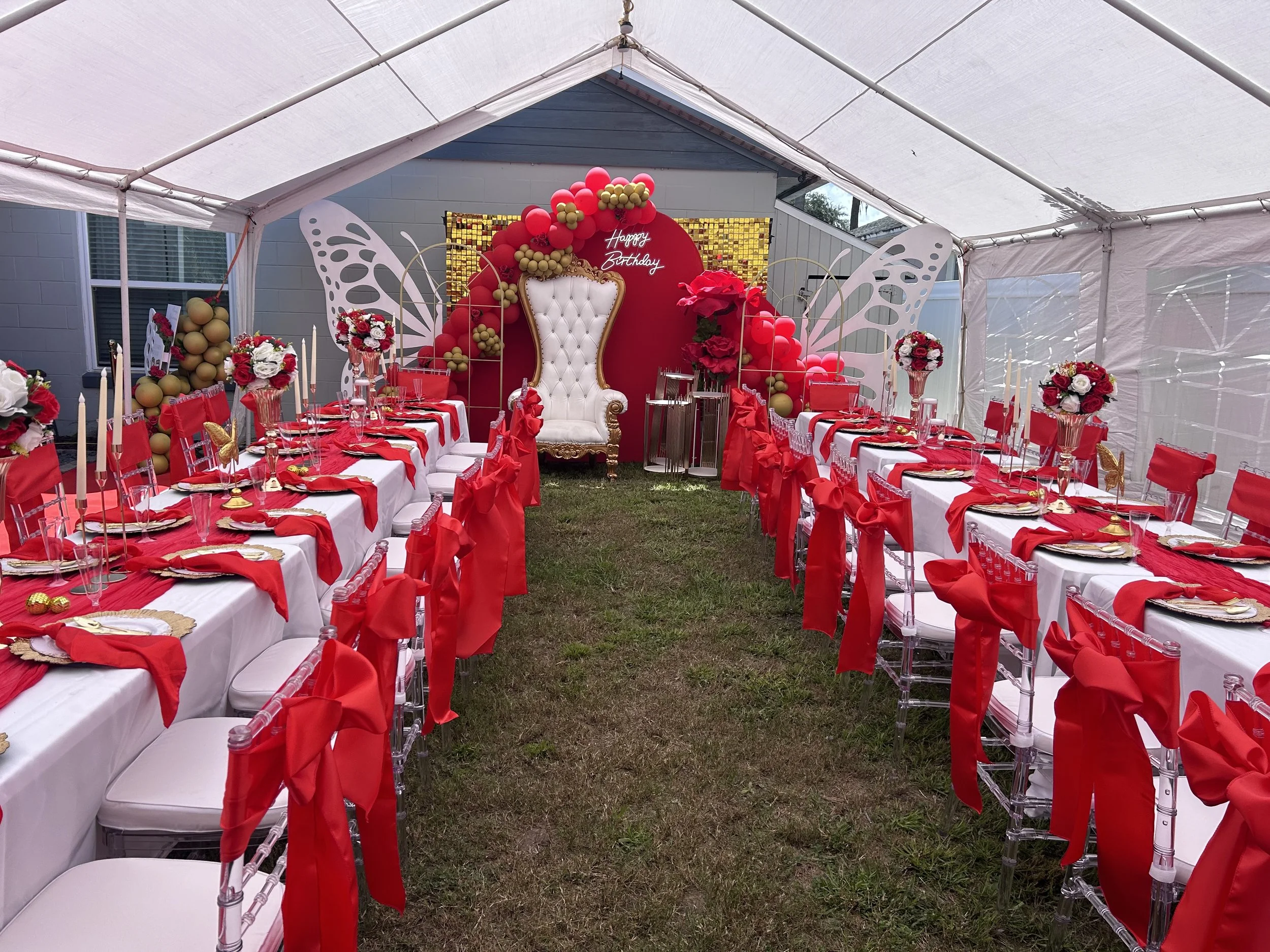 Decorated outdoor birthday celebration setup with a white tent, long tables with red and white table settings, floral centerpieces, and a backdrop featuring a white throne with a red floral arch and a "Happy Birthday" sign, surrounded by butterfly wing decorations.