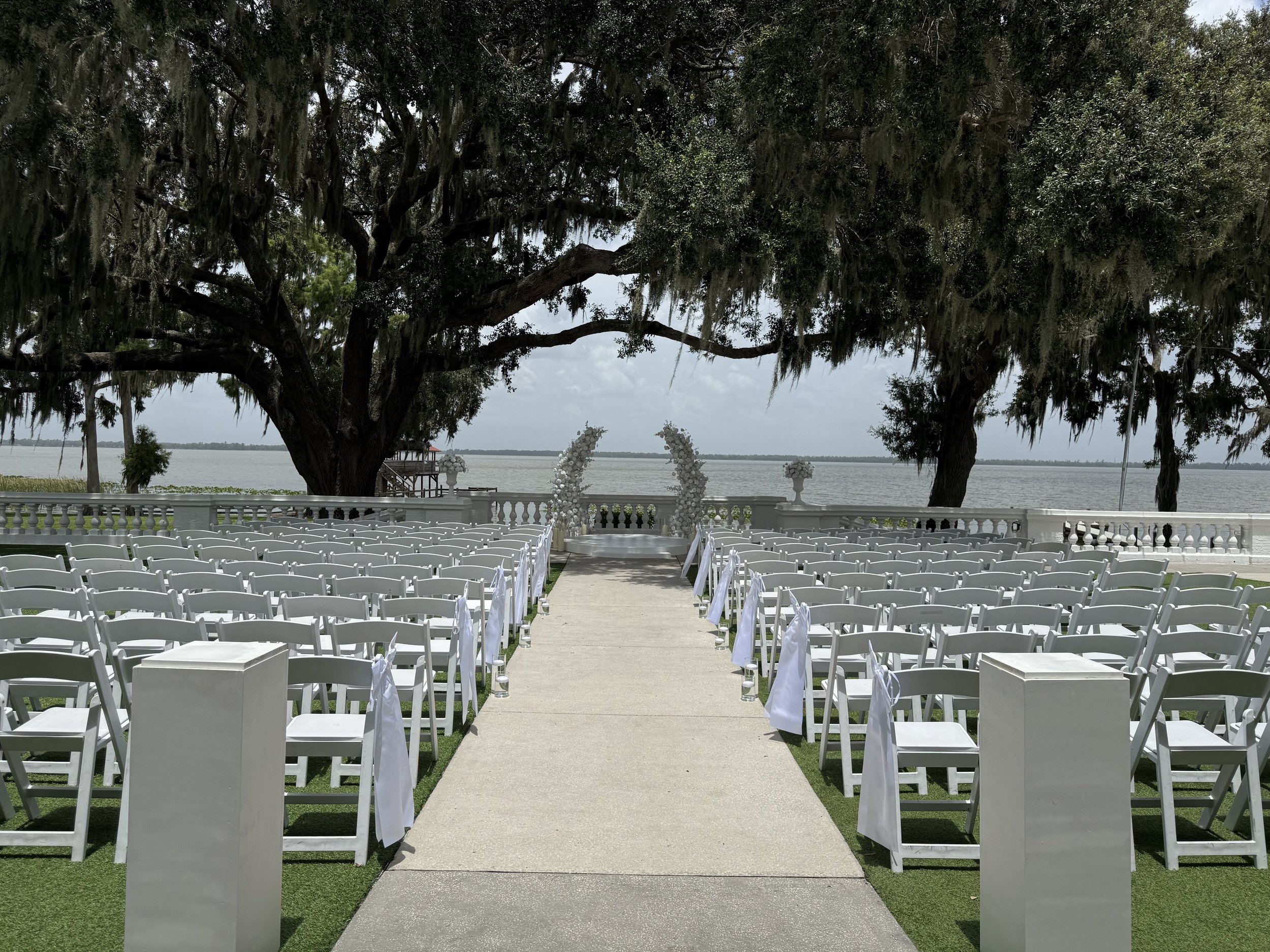 Outdoor wedding setup with rows of white chairs along a central aisle leading to an arch of white flowers, with large trees and a body of water in the background.