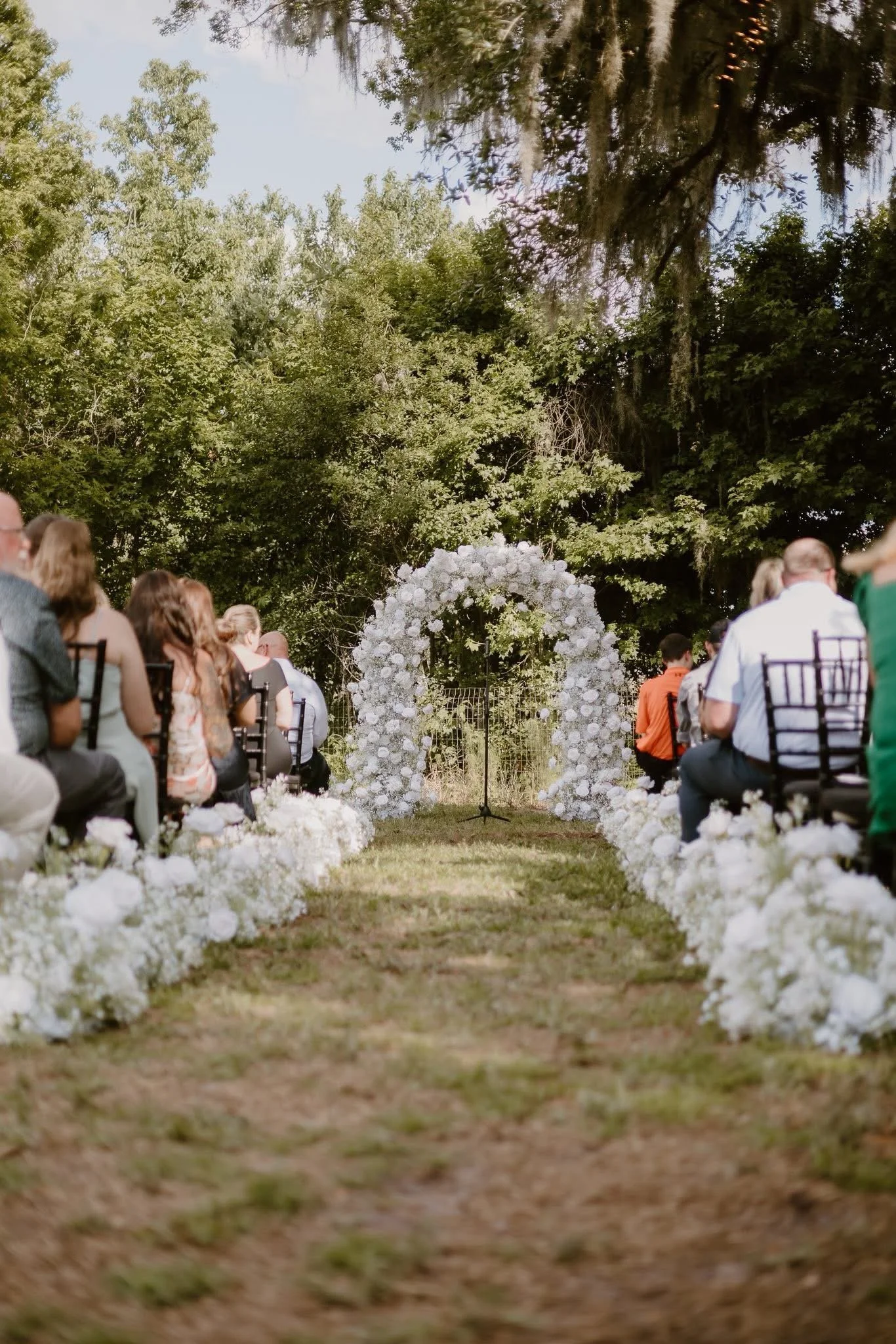 Outdoor wedding ceremony setup with flower arch, chairs, and guests seated on both sides of the grassy aisle, surrounded by trees.