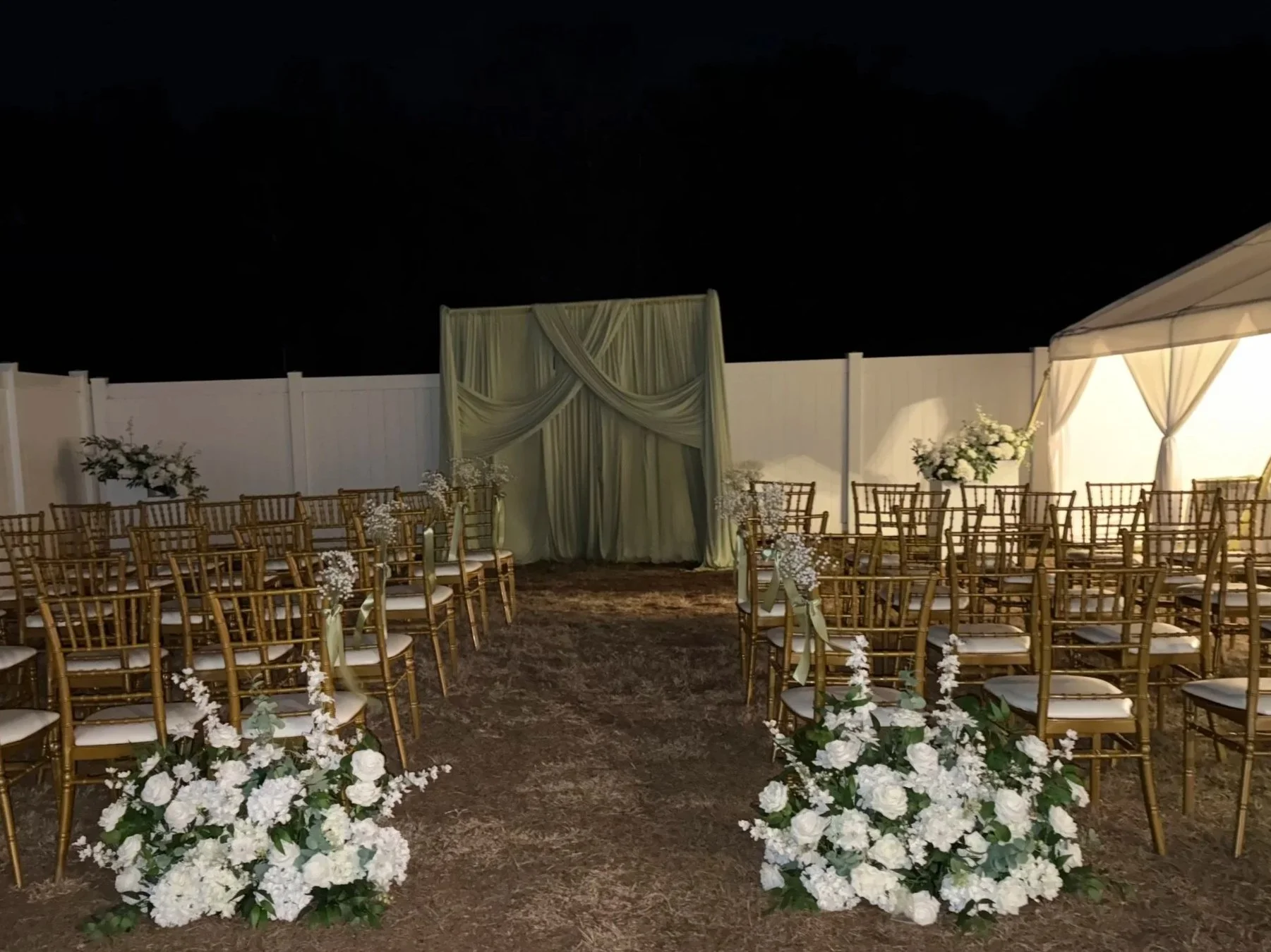 Outdoor wedding setup at night with golden chairs arranged in rows facing an altar with draped white fabric and floral arrangements. Flower decorations line the aisle, and a white fence surrounds the area.