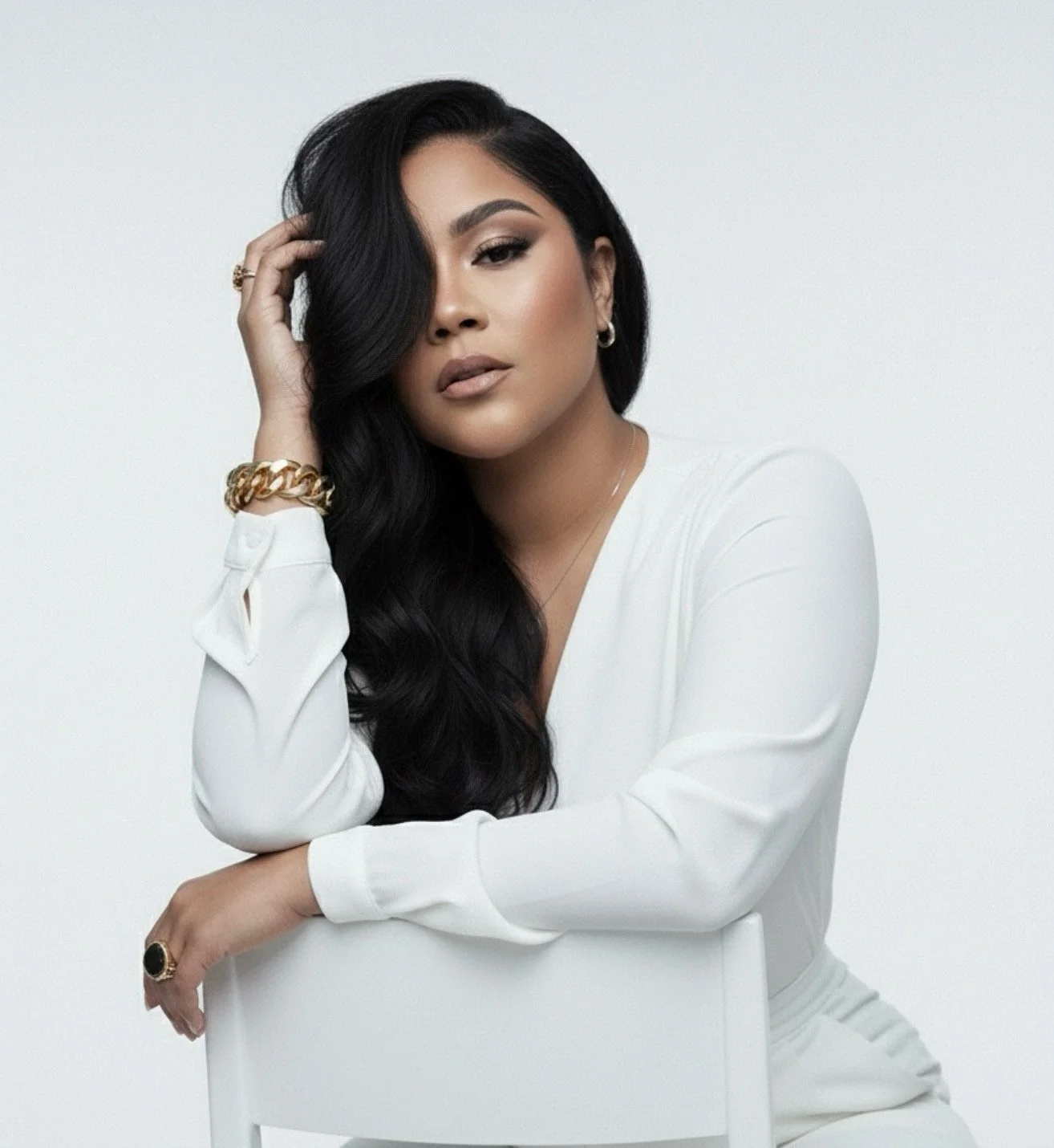 A woman with long black hair styled in loose waves, wearing a white long-sleeved top and gold jewelry, poses with her hand touching her hair against a plain white background.