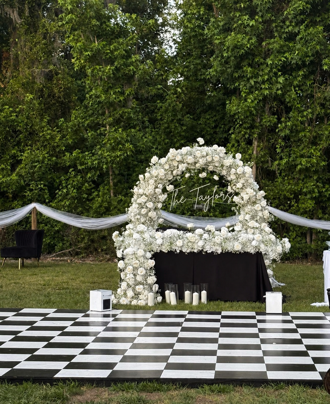 Outdoor wedding or celebration setup featuring a black and white checkered dance floor, a large floral arch with white roses and greenery, a sign that says 'The Taylors,' candles in glass holders, and a background of lush green trees.