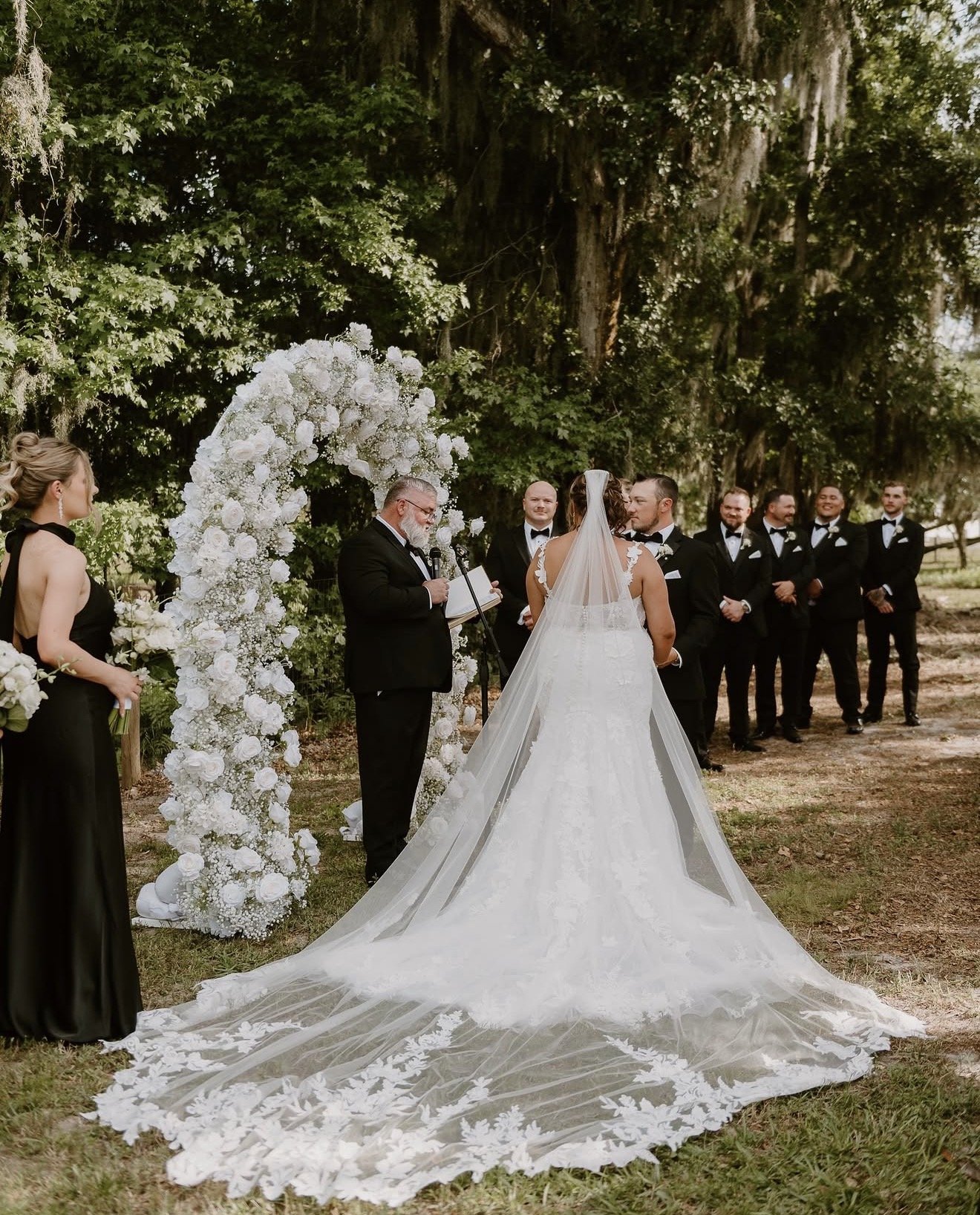 A outdoor wedding ceremony with a bride and groom, officiant, bridesmaid, and groomsmen, with a floral arch and lush green trees in the background.