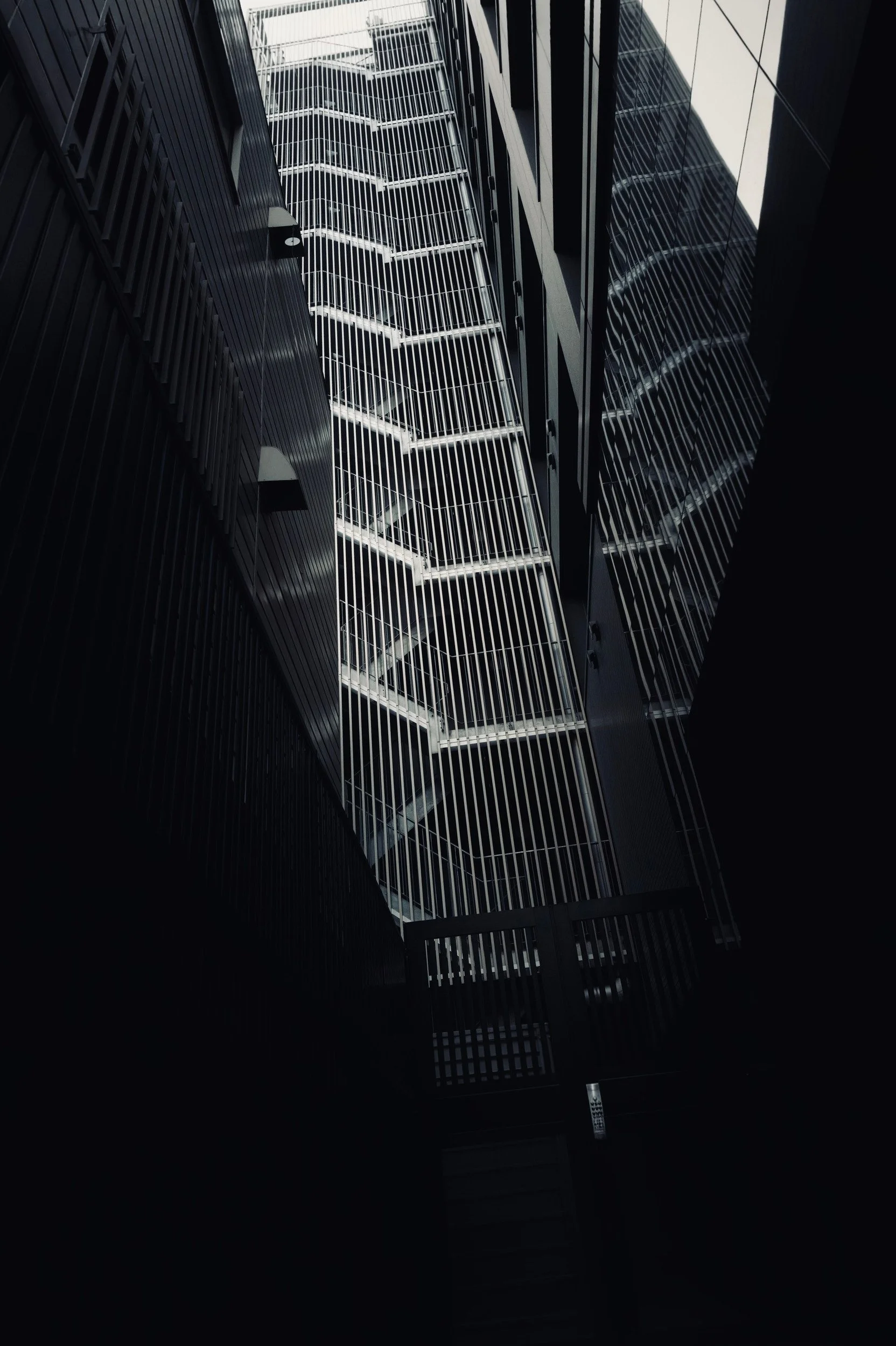 An exterior metal staircase suspended between dark facades, with a reflection in adjacent glass.
