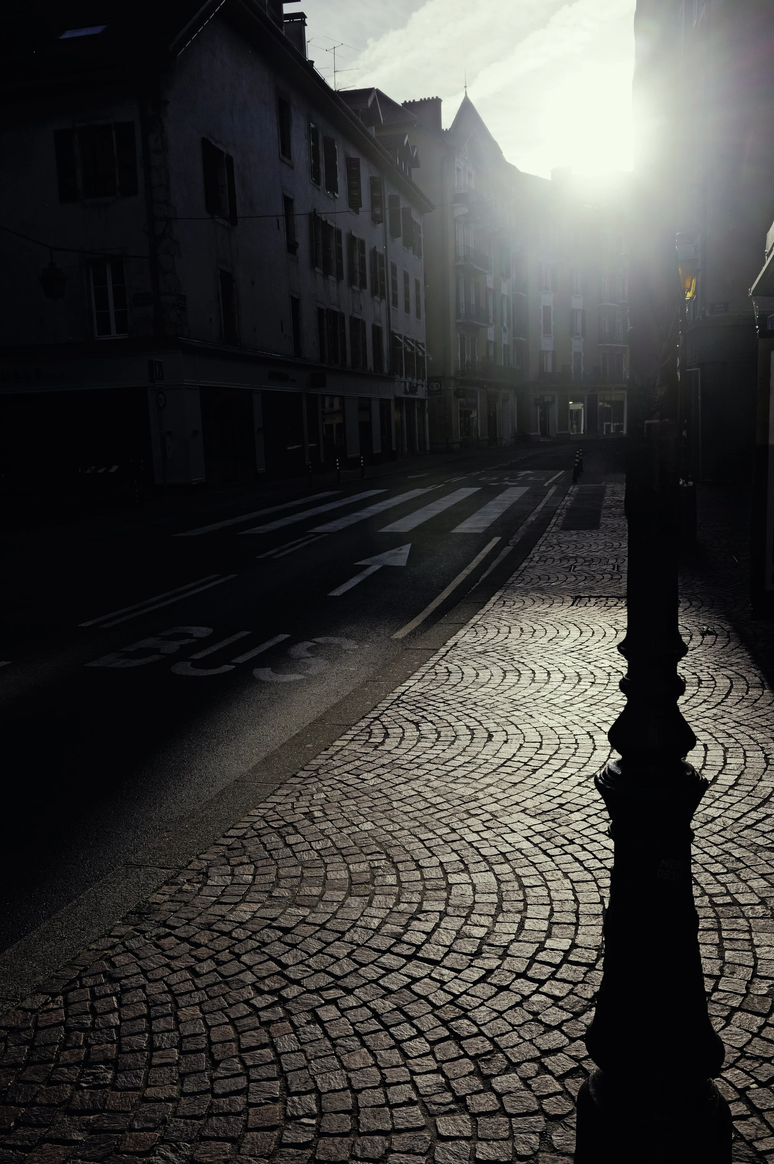 High-contrast urban photography: the geometric shadow of a street lamp cast across a cobblestone road.