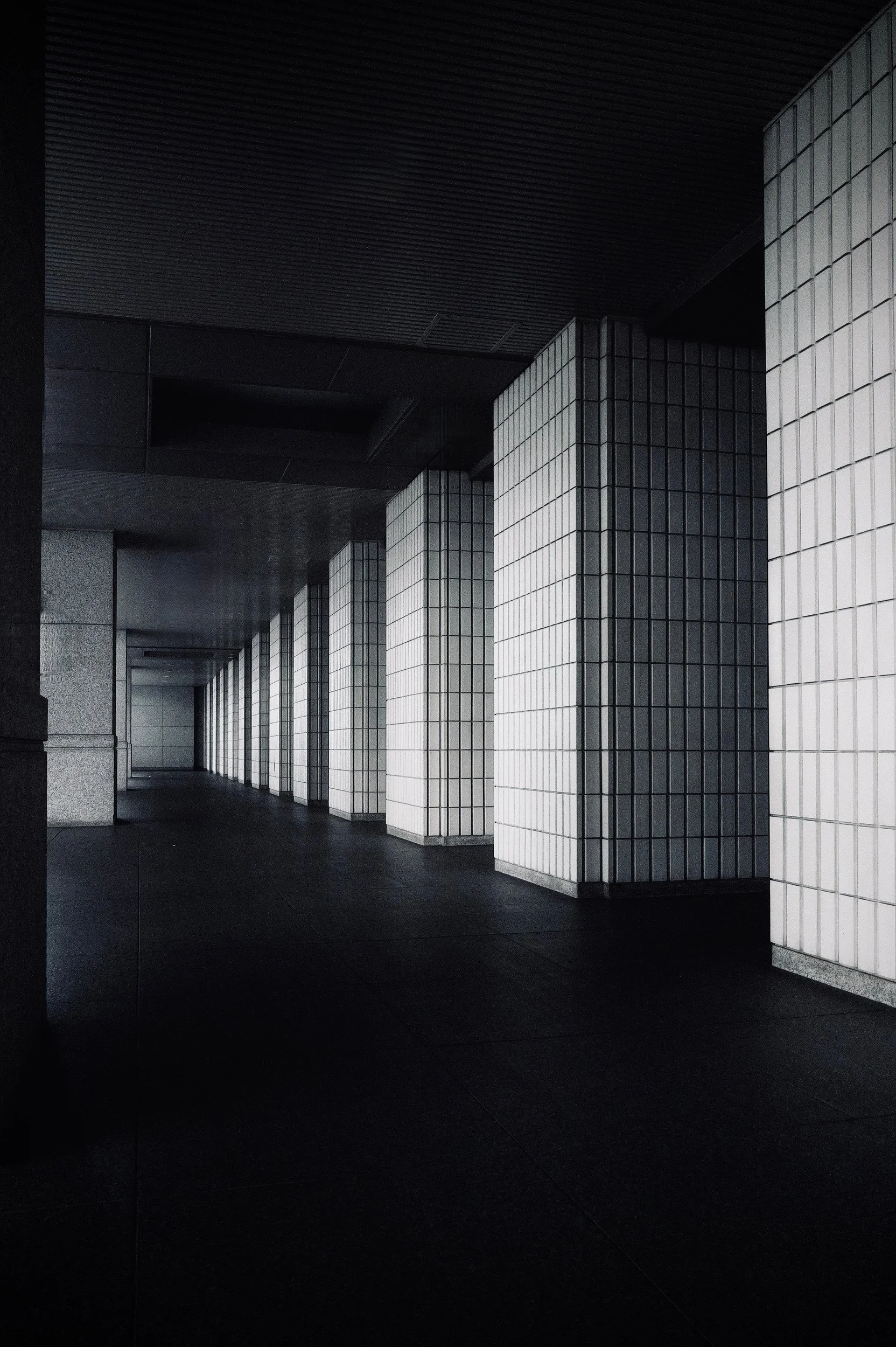 A deserted covered corridor with repeating square tiled columns and a dark floor.