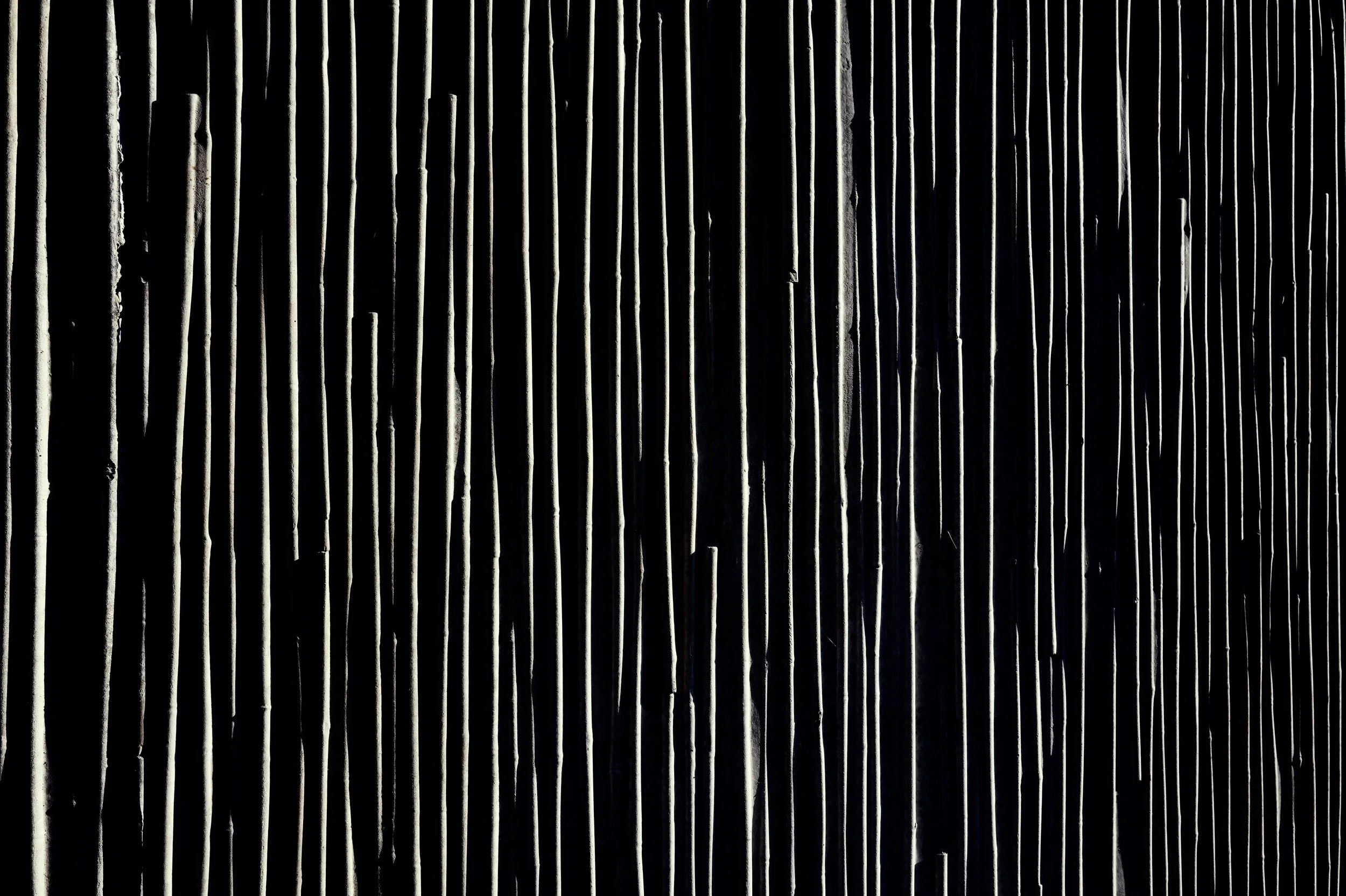 Textured close-up of an architectural structure with repetitive vertical lines. Extreme contrast between deep black shadow and white light, urban minimalist photography aesthetics.