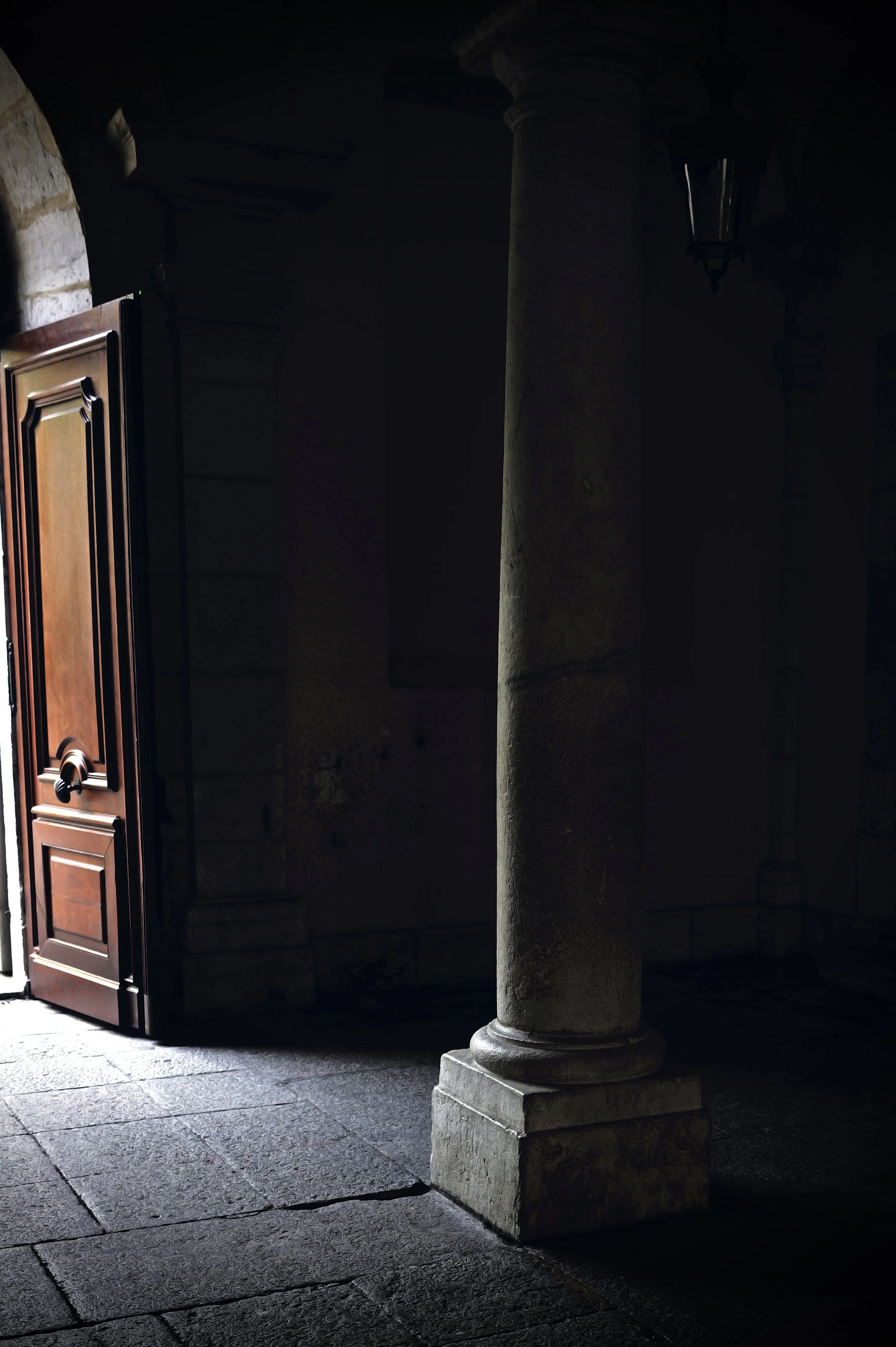 Interior architectural photography. Dramatic light entering through a wooden door onto stone floors. High contrast and silence.