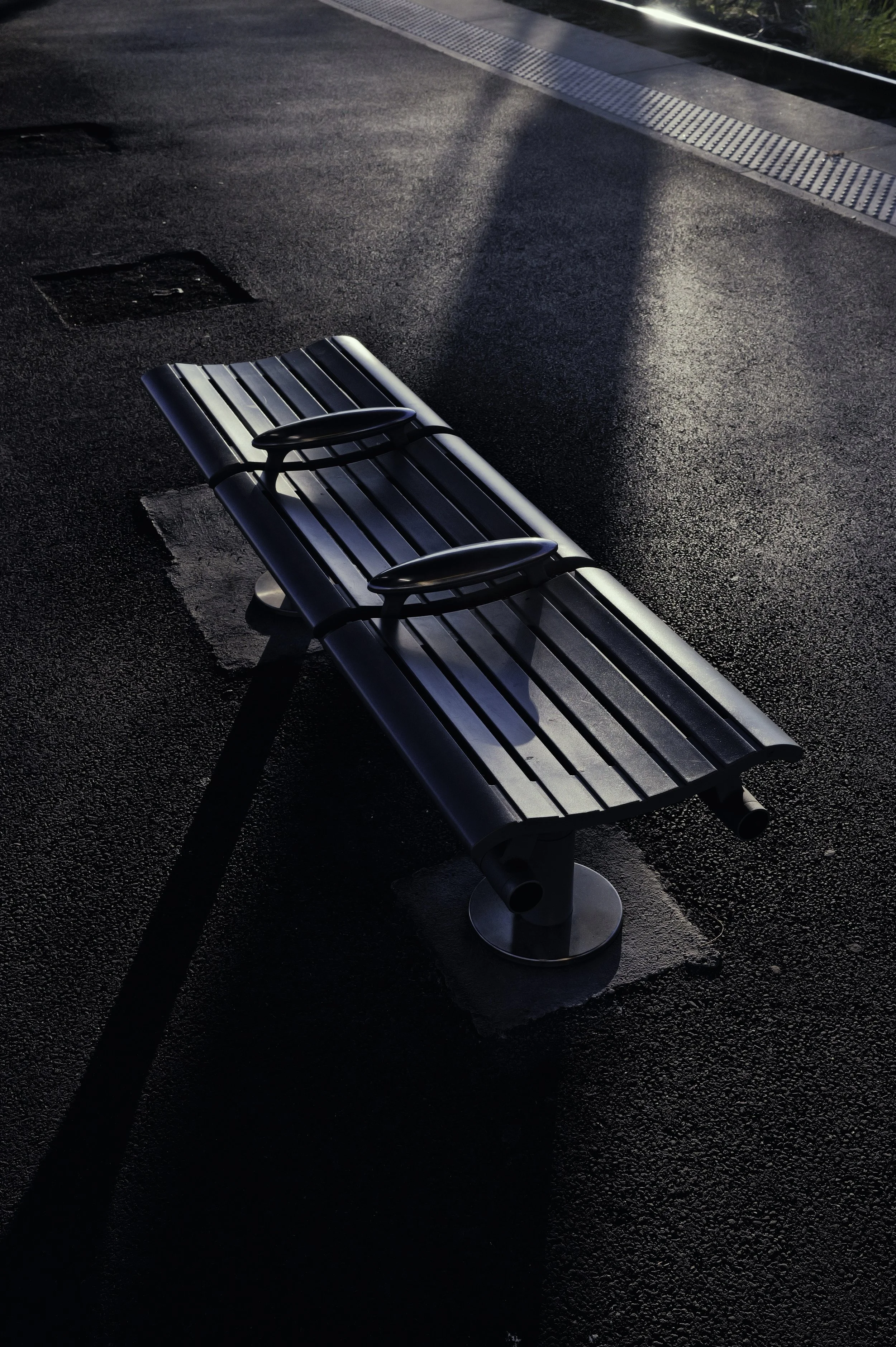 Minimalist urban detail of a metal bench on asphalt with long sunlight shadows. Urban Chrome street photography.